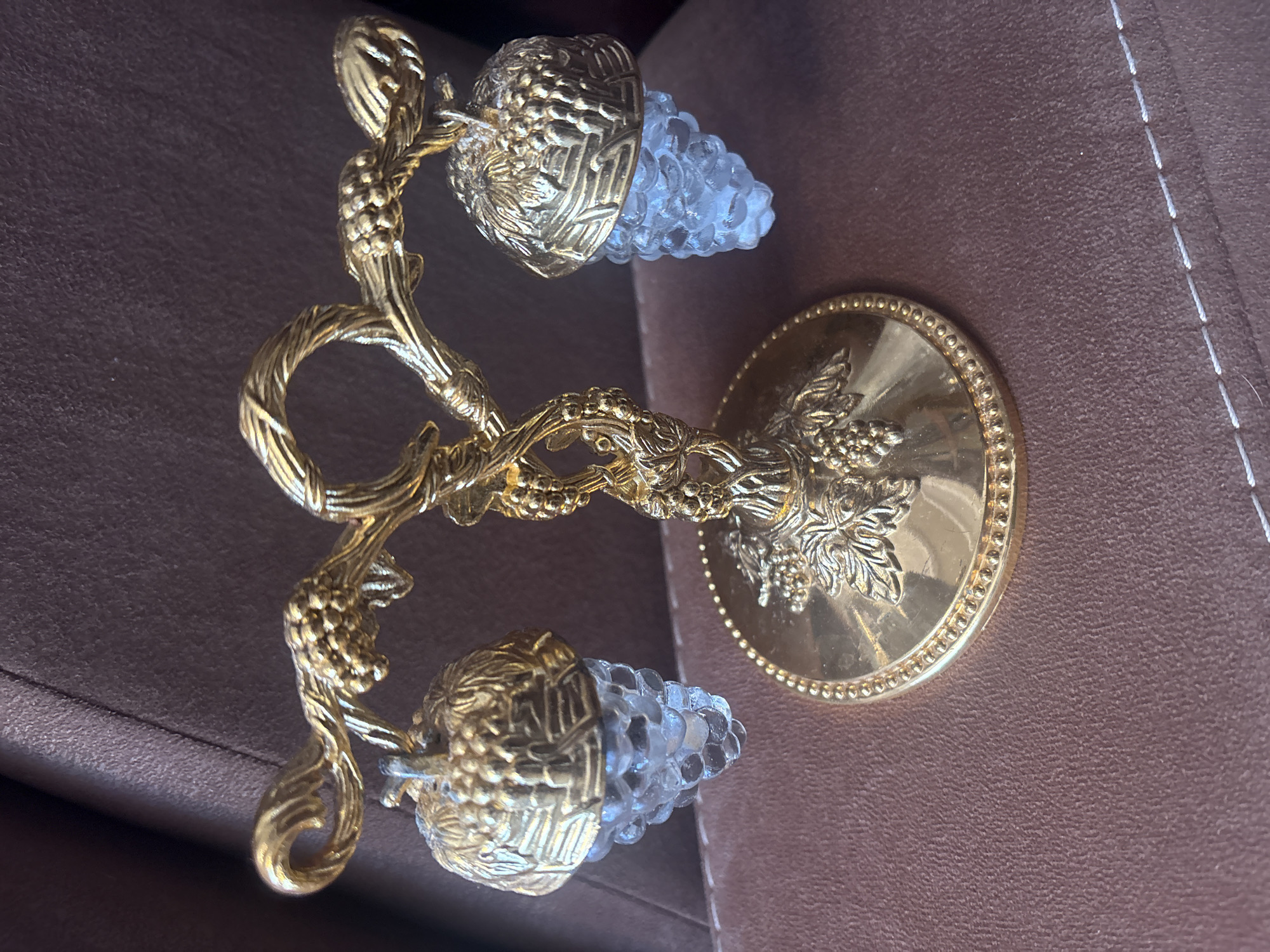 A decorative, golden-colored metal candelabra with two arms holding glass grape bunches is photographed from above on a brown, textured surface. The candelabra features a central stem sculpted to resemble intertwined grape vines and leaves, with each arm extending to hold a glass bunch of grapes. The base is a circular gold-colored disc with a grape and leaf motif. The lighting is bright, highlighting the metallic sheen of the candelabra. The texture of the surface suggests it could be fabric or leather. There is no indication of the location being Krasnoyarsk, Russia.