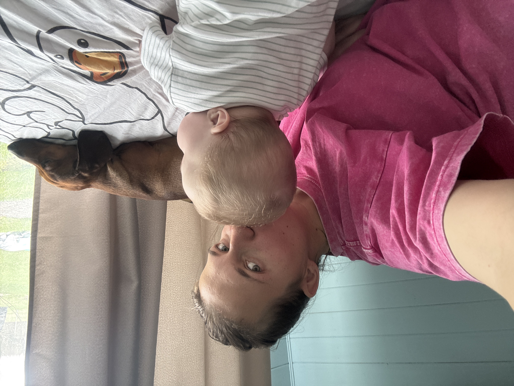 An intimate indoor scene features a woman, a baby, and a dog. The image is oriented sideways; if rotated clockwise, it shows a woman lying down, looking directly at the camera with a focused expression. Her head is positioned to the right, with a baby's head close to her face, suggesting a moment of affection or closeness. The baby, with light blonde hair, is wearing a white top with thin grey stripes. The woman is dressed in a textured, pink short-sleeved t-shirt.
To the left of the baby, a brown dachshund dog is resting on a white duvet adorned with a black outline drawing of a duck. The background reveals a bright window on the far left, allowing daylight to illuminate the scene and showing greenery outside. Grey curtains partially cover the window. On the right side of the frame, behind the woman, there is a light blue wall with horizontal paneling. The overall atmosphere conveys a peaceful, shared moment among family members and their pet. No visible text is present in the image.