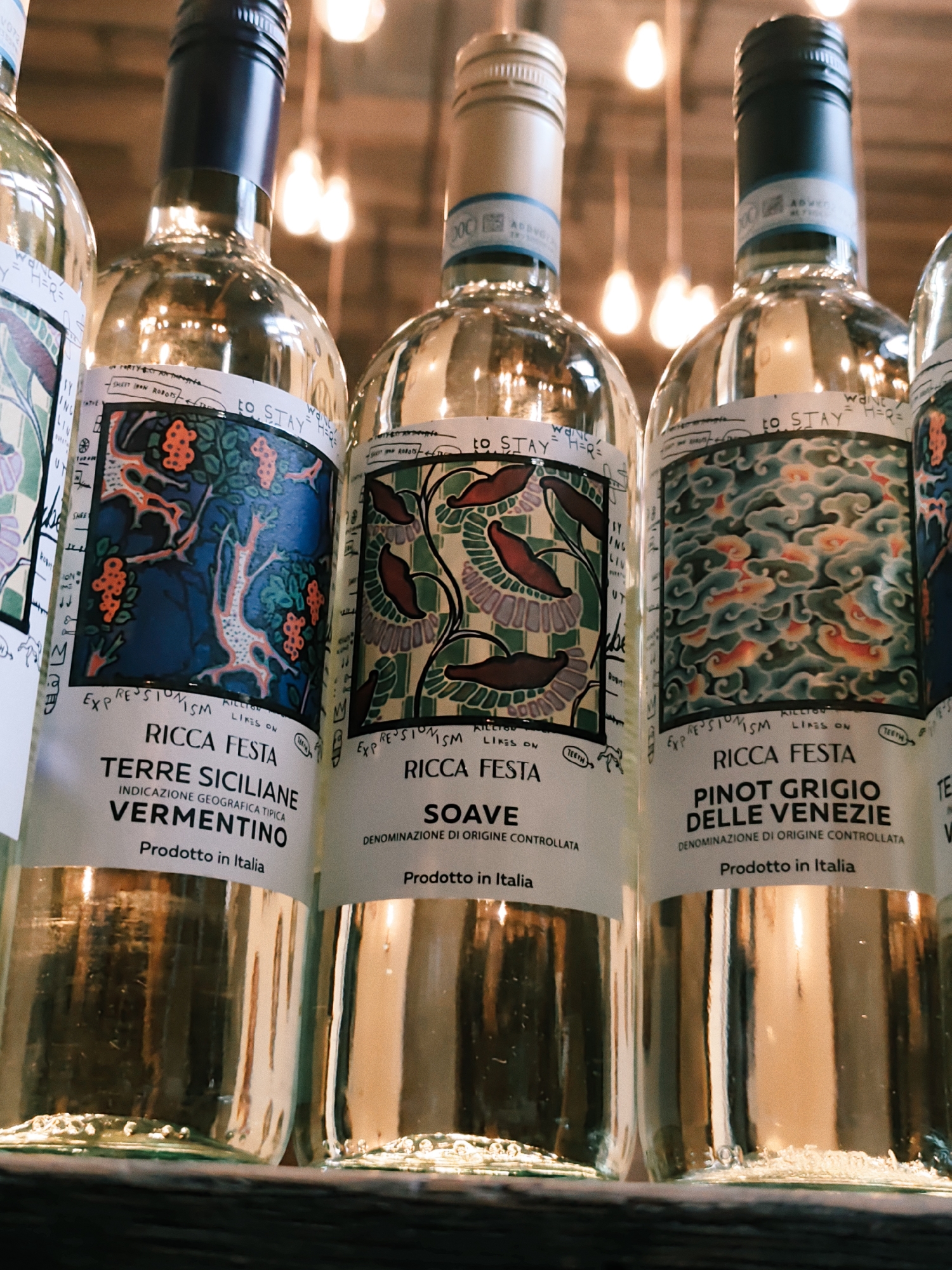 This is a close-up, eye-level shot of several bottles of white wine lined up on a shelf. The bottles are made of clear glass and are filled with a pale yellow liquid. Each bottle has a unique label with an artistic design and text.
The labels indicate that the wines are "RICCA FESTA" brand, with varieties including "TERRE SICILIANE VERMENTINO," "SOAVE," and "PINOT GRIGIO DELLE VENEZIE." The text also mentions "INDICAZIONE GEOGRAFICA TIPICA" and "DENOMINAZIONE DI ORIGINE CONTROLLATA," suggesting they are Italian wines. Some labels have handwritten elements, such as "to STAY" and drawings. The bottles are arranged closely together, and the background is softly blurred, showing warm, out-of-focus lights, possibly from lamps or string lights, creating a cozy and inviting atmosphere. The lighting in the scene is warm, suggesting it might be evening or an indoor setting. The focus is sharp on the labels and the upper parts of the bottles, while the bottoms of the bottles are slightly blurred, hinting at a shelf or surface they are resting on. There are no people visible in the image. The scene appears to be a wine shop, cellar, or a bar setting.