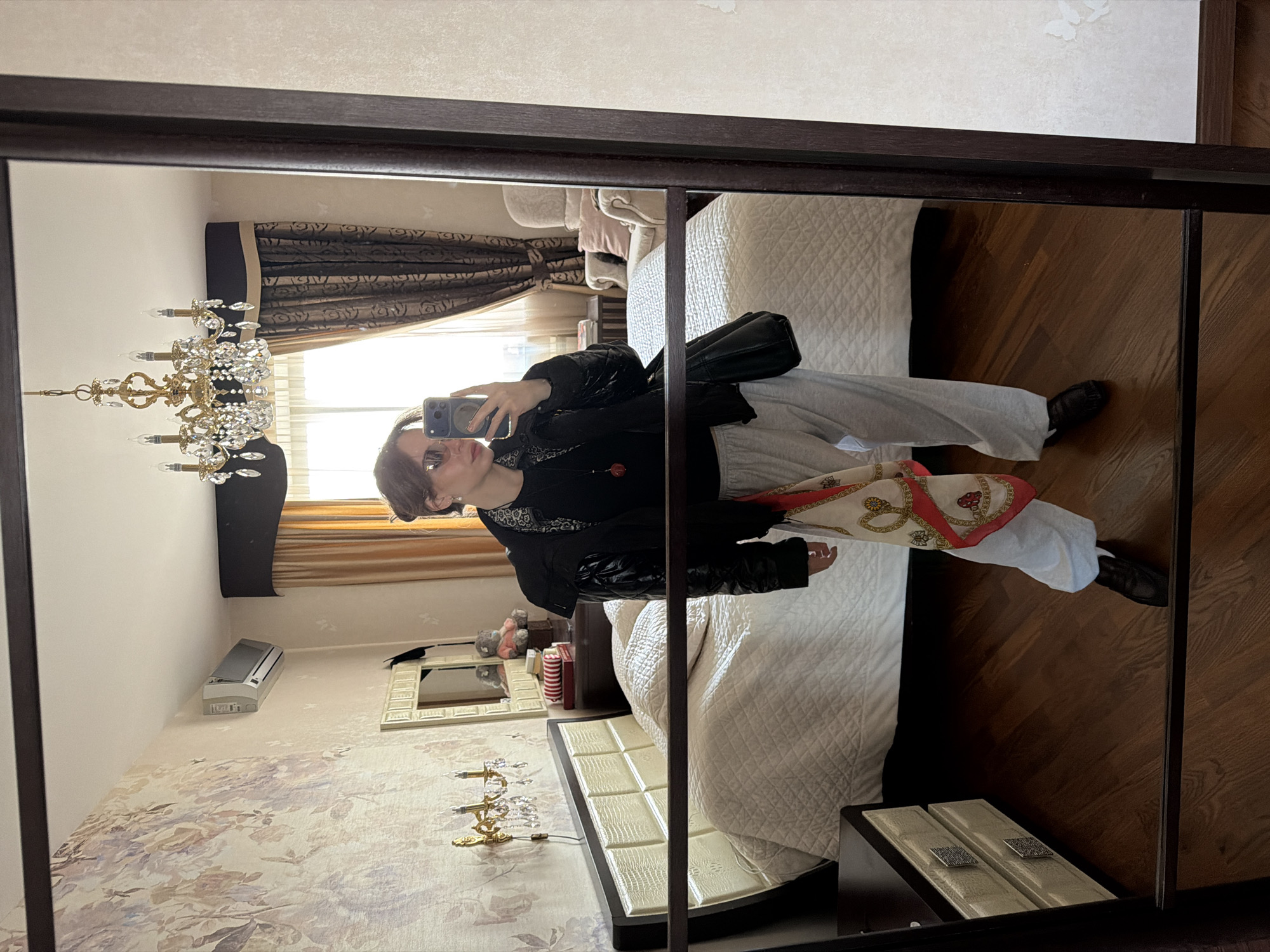 A person is reflected in a large mirror within a room. The mirror has a dark brown frame. The person, likely taking a selfie, is wearing a dark jacket, light-colored pants, and dark shoes. They are also holding a black bag.
The room reflected in the mirror features a bed with a white quilted bedspread. There is a chandelier visible in the reflection and curtains over the window. Additional details within the room include wallpaper with a floral pattern, an air conditioning unit mounted on the wall, and a vanity table.
The location is Khimki, Russia.