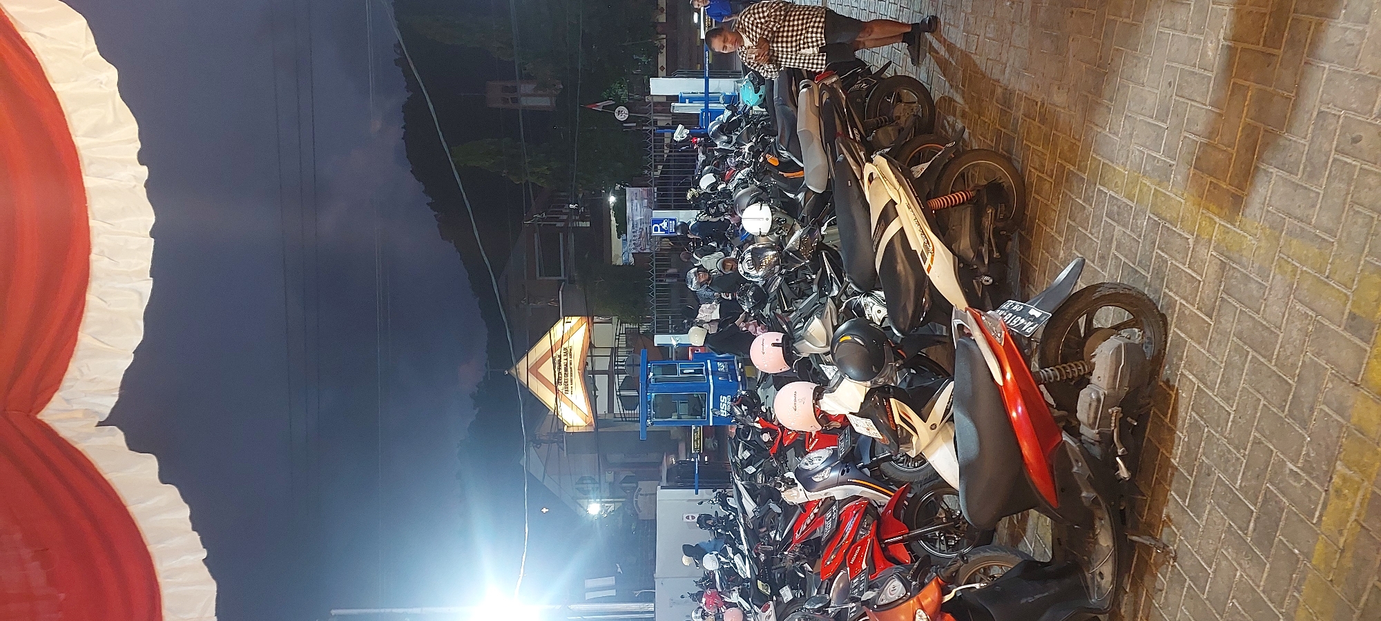 This is a photograph of a motorcycle parking area in front of a building in Jayapura, Indonesia, at night. The foreground is dominated by a tightly packed array of parked scooters and motorcycles. Several helmets, including pink and black ones, are visible atop the parked vehicles. The pavement is made of light-colored interlocking bricks with yellow painted lines.
In the mid-ground, a blue ticket booth for parking is situated, with "BSS PARKING" signage. Behind the ticket booth, a building with a triangular, illuminated facade is visible. Text on the building's facade reads "GEREJA KANAL RESORT & SAIN" and what appears to be "PIROMG. REGEI & SAIN". This likely indicates the building is a church or religious institution. A tall, bright light pole illuminates the area from the left.
In the background, a dark, tree-covered hillside rises, with a few buildings and structures visible on its slopes. The sky is dark, indicating nighttime. A woman in a checkered shirt and shorts stands to the right of the ticket booth, holding a phone to her face. Several other people, mostly wearing helmets, are seen near the parked motorcycles. Red and white bunting is draped across the top of the image, suggesting a festive occasion.