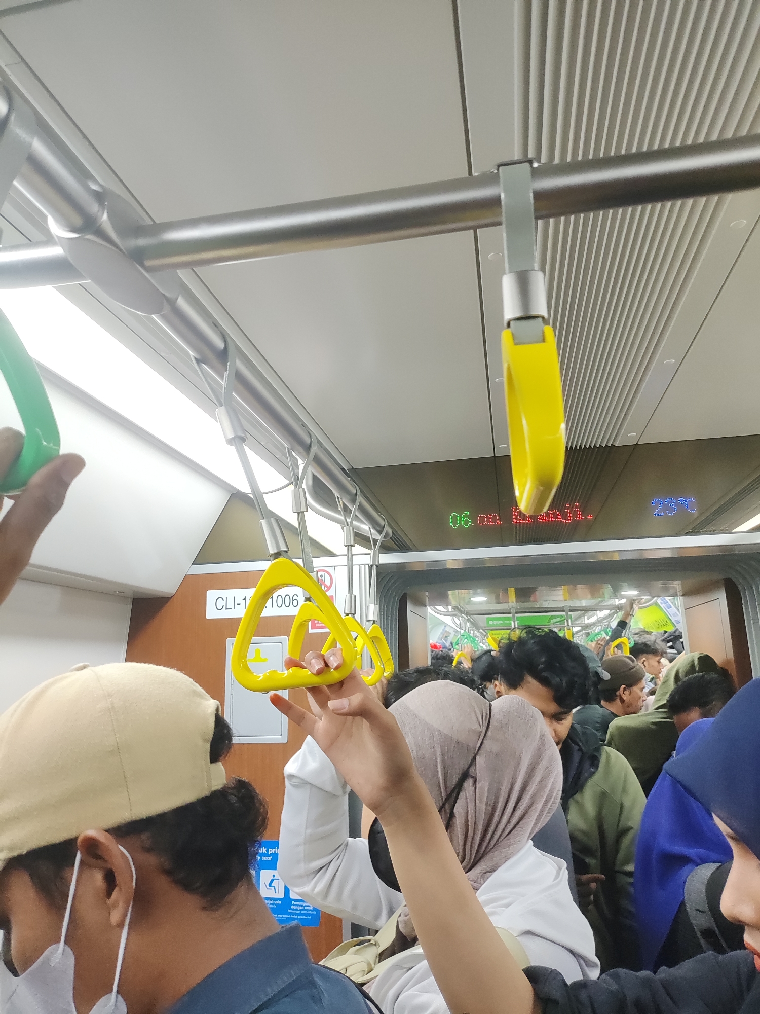 An indoor photograph captures a crowded subway car with passengers holding onto yellow and green handrails. The car interior features metal poles and ribbed ceiling panels. A digital display above the windows shows "06:00 Km.1006" and "23°C". A passenger in the foreground wears a beige cap and a white face mask. Another passenger in the mid-ground is dressed in a white long-sleeved shirt and a tan headscarf. The lighting is bright and overhead.