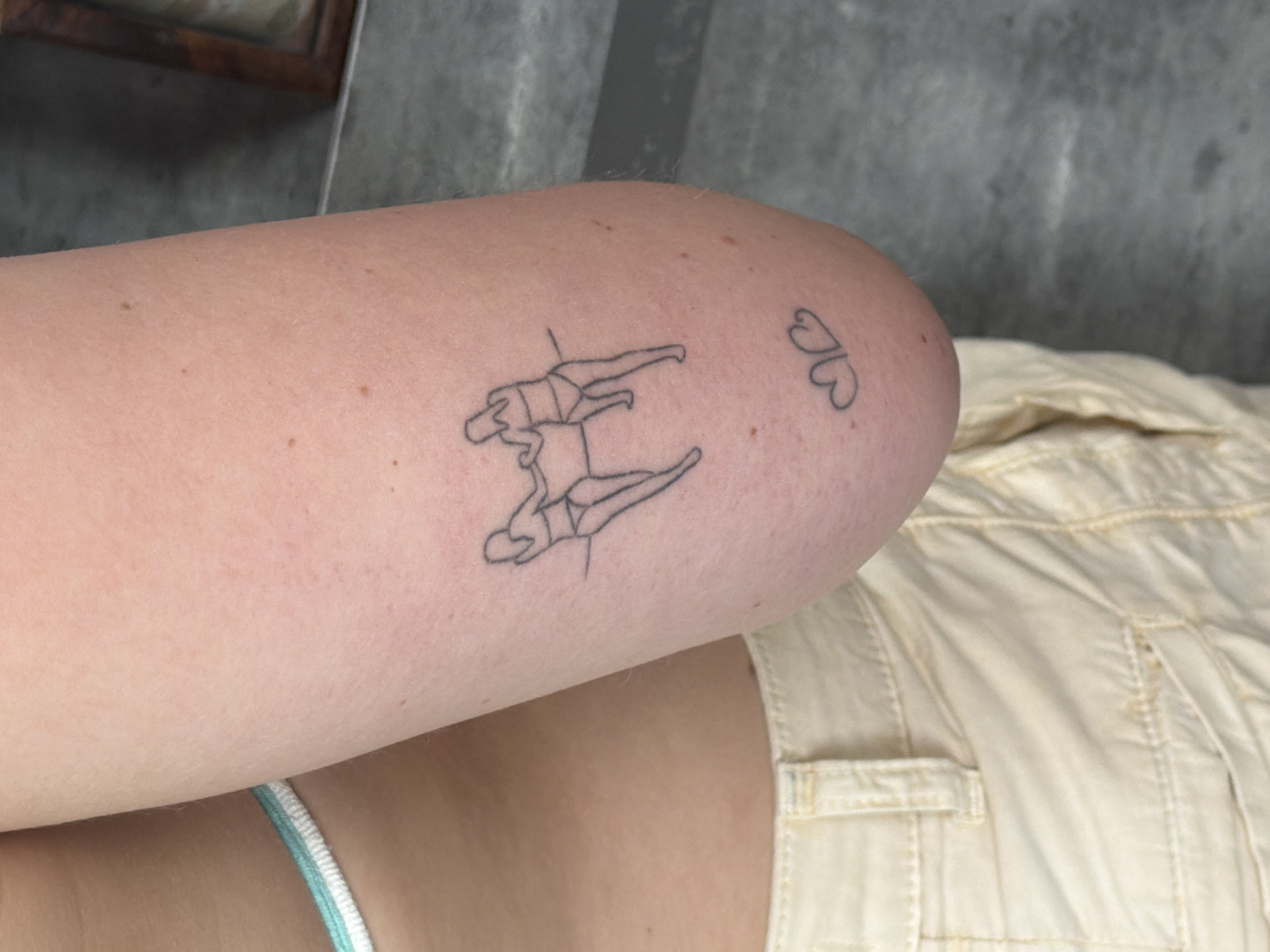A close-up shot depicts a person's arm, presumably the left arm, adorned with two tattoos. One tattoo features two mirrored stick-figure women balancing on a pole. The second tattoo located on the upper arm consists of two small hearts that are side-by-side. The person is wearing beige clothing and a blue-striped garment is visible near their torso. The background is blurred. The image does not appear to contain visual information that would allow the location to be determined.