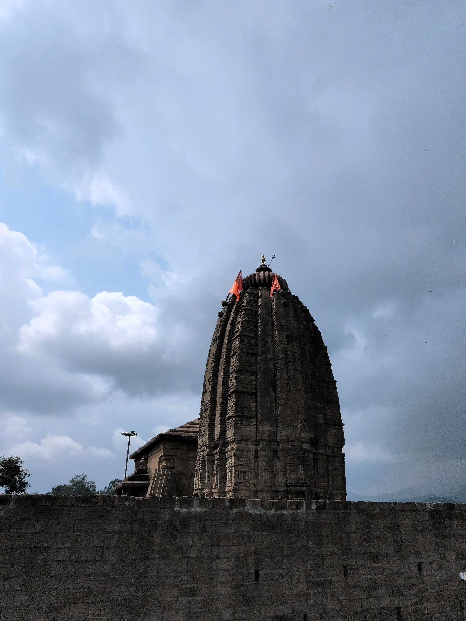 This image captures a monumental stone temple structure with a tiered, conical roof against a dramatic, overcast sky. Two saffron-colored flags are prominently displayed at the peak of the temple, adding a vibrant contrast to the muted tones of the architecture and sky. The temple is built with intricately carved stone blocks, showcasing detailed craftsmanship. A stone wall in the foreground forms a barrier, separating the viewer from the temple grounds. The background reveals hints of distant hills and trees, suggesting a rural or semi-rural setting. The overall atmosphere is one of solemnity and grandeur, possibly on a day with impending rain. The location is identified as the temple of Baijnath, in Baijnath, India. There are no people or discernible actions visible in the image.