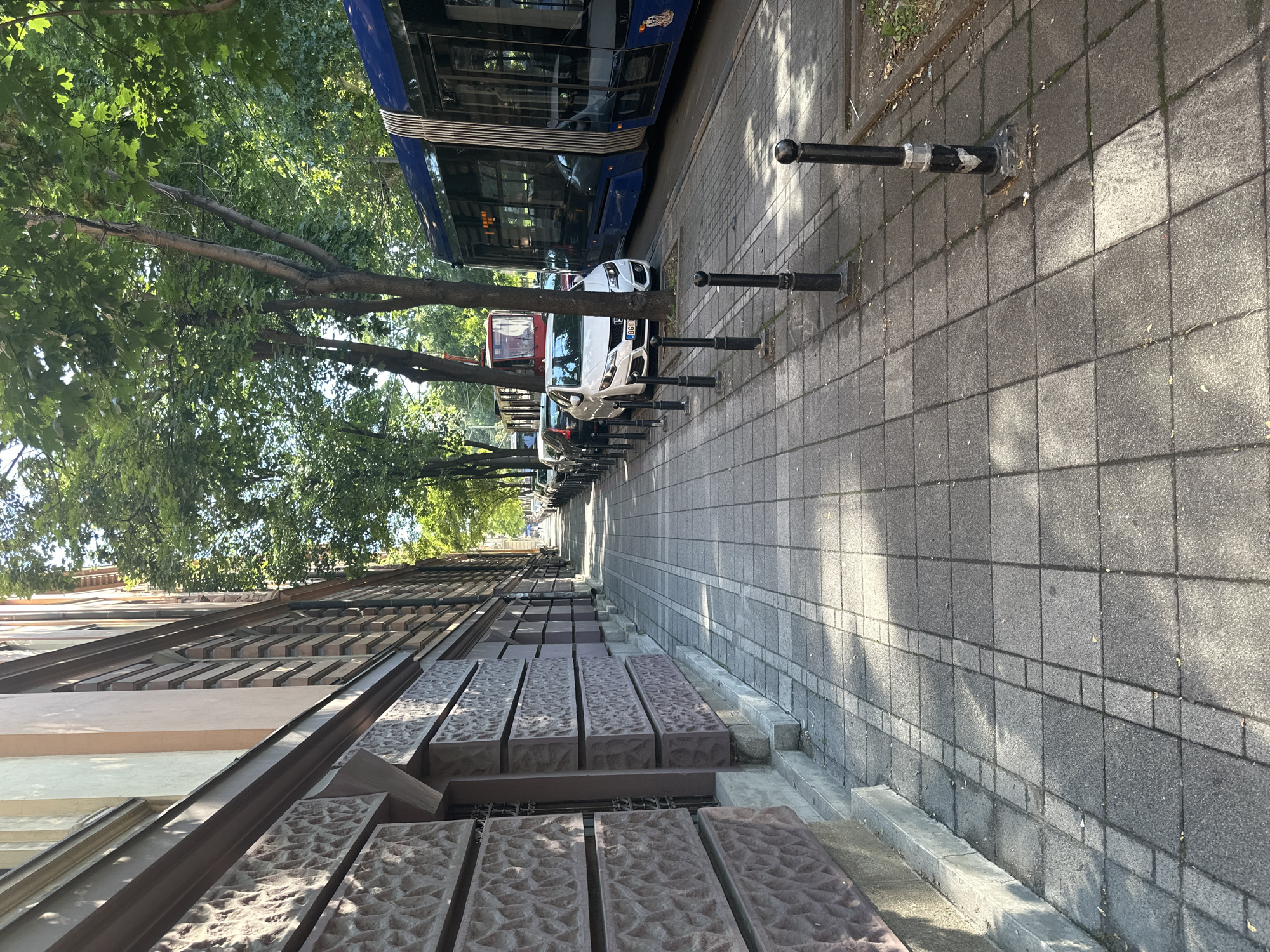 The image depicts a sun-drenched sidewalk in Belgrade, Serbia, adjacent to a building with an ornate stone facade. The sidewalk is paved with rectangular tiles and lined with black bollards. A modern blue tram is visible on the right, with its doors open. Several cars are parked along the street, and trees with lush green foliage cast dappled shadows on the walkway. In the background, more buildings and trees suggest an urban environment. The scene is static, with no discernible human activity or interactions.