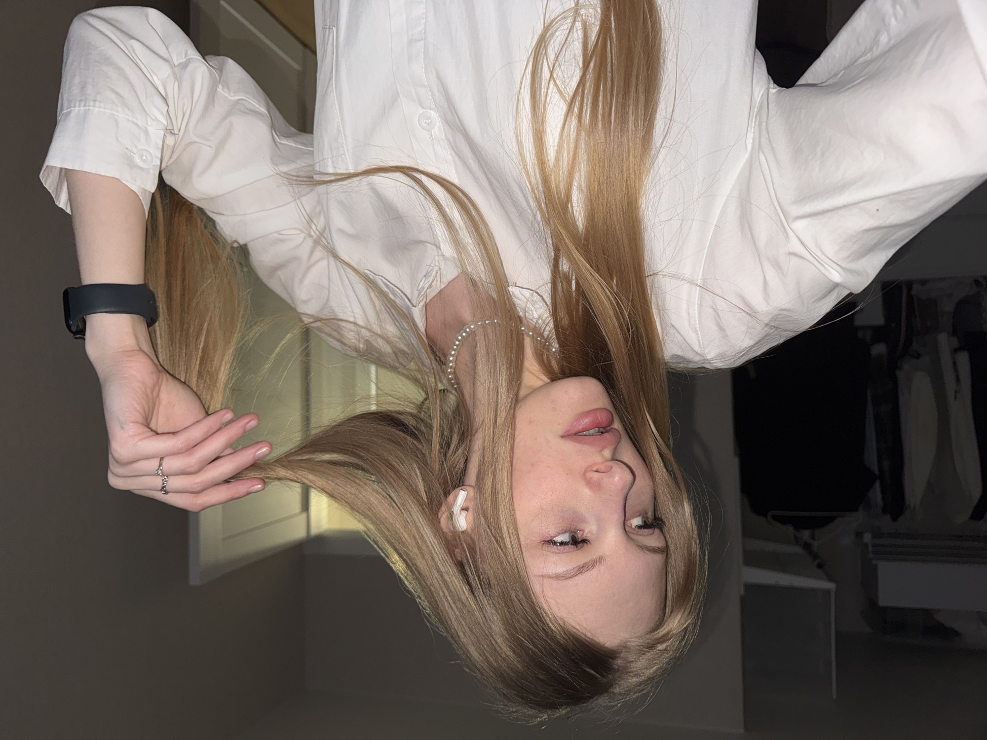 A young woman with long, light brown hair and fair skin is depicted in a close-up, chest-up shot, angled as if looking up and to the right, with her head slightly tilted. She wears a crisp white collared shirt and a delicate pearl necklace. A white wireless earbud is visible in her right ear. Her left hand, adorned with a silver-colored ring on the ring finger, gently holds a section of her hair. A black fitness tracker or smartwatch is visible on her left wrist. Her lips are slightly parted, and her expression is neutral and somewhat contemplative. The background is mostly dark and out of focus, indicating an indoor setting, possibly a room. The lighting appears soft and somewhat dim. No text is visible in the image. The location context provided is Perm, Russia.