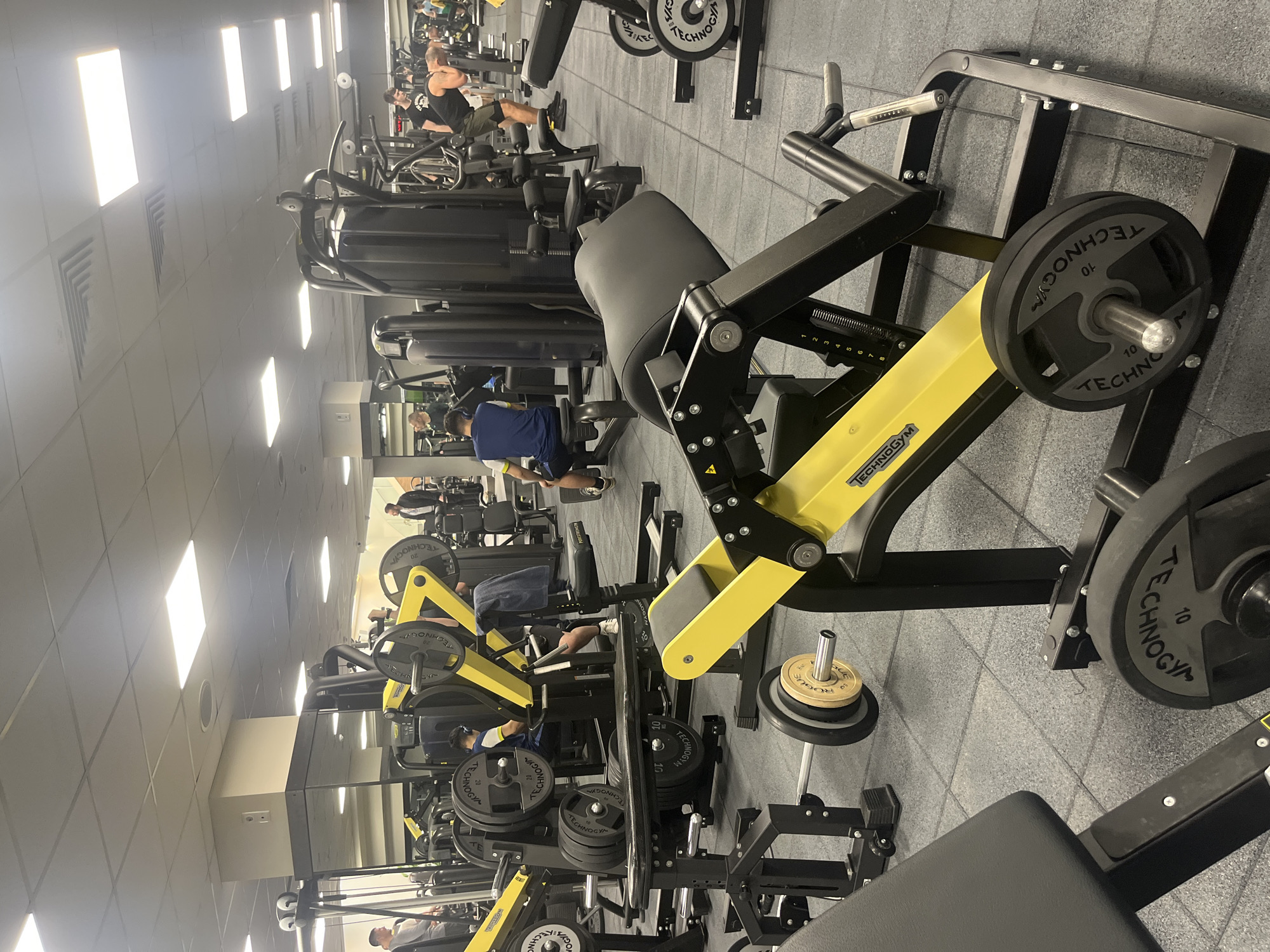The image shows the interior of a fitness center. The gym is well-lit with multiple overhead lights, and the floor is covered with gray tiles. The gym has several weight machines with black frames and yellow accents, with some machines loaded with weight plates. There are people using the equipment in the background. The focus is on a selection of leg press machines. The gym machines bear the "Technogym" logo. Specific city location is unavailable.