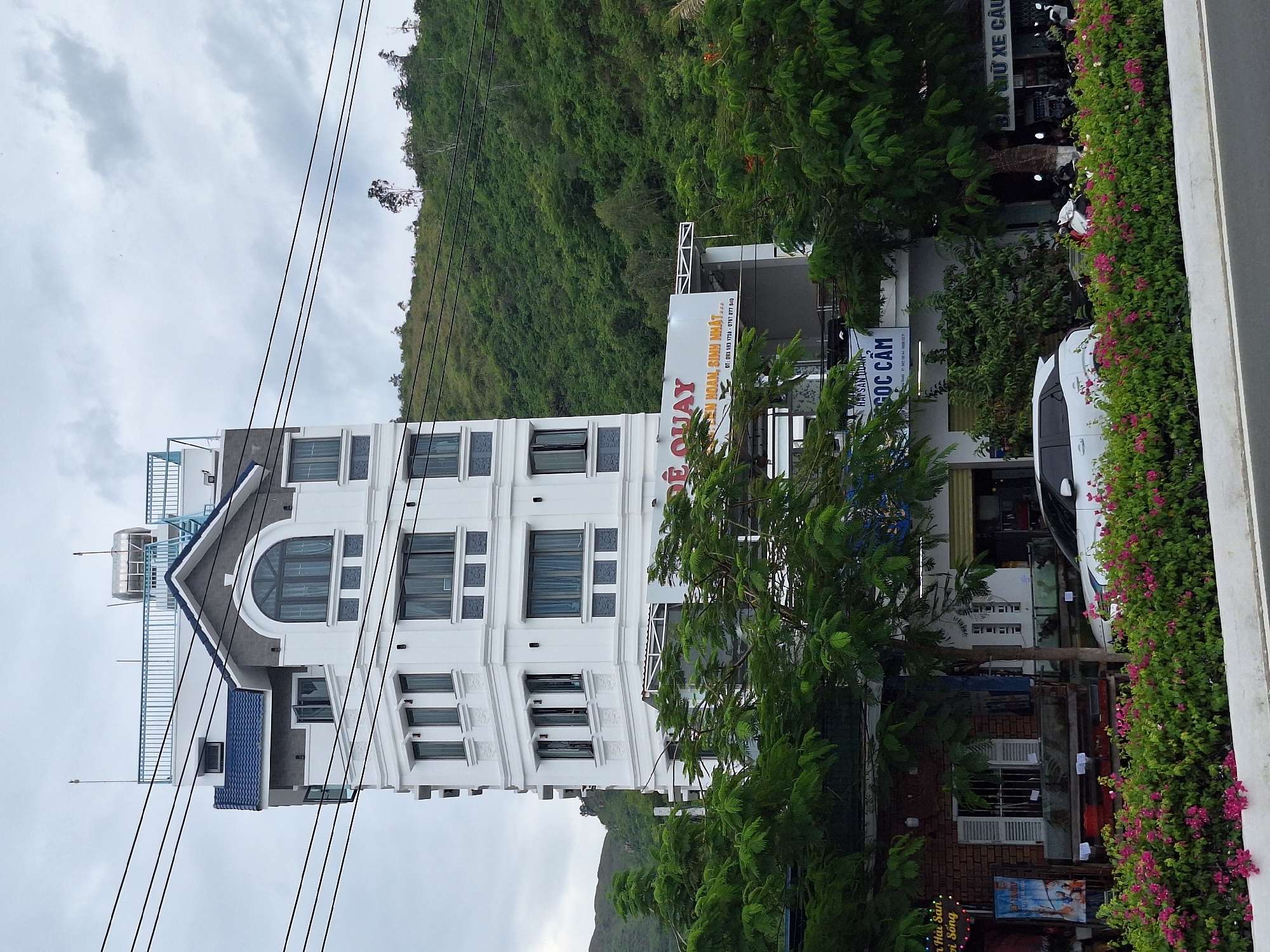 The image depicts a multi-story white building with dark blue accents, likely a hotel or apartment complex, situated in Nha Trang, Vietnam. The building features multiple windows and decorative architectural elements. In front of the building, there are lush green trees and a vibrant hedge with pink flowers, suggesting a roadside setting. Power lines are visible crisscrossing the sky. Behind the building, a steep, tree-covered hill rises. A white car is parked in front of the building, and several motorcycles are parked further down the street. There are signs on the building, one of which reads "ĐỂ QUAY" and another "NGỌC CẨM". The sky is overcast.