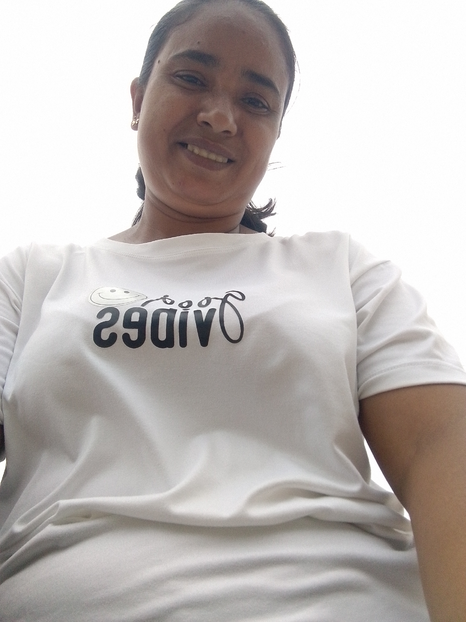 A woman is photographed from a low angle. She is smiling and wearing a white t-shirt with "Vibes" printed backwards. The background is a bright, overcast sky. The location of the photograph is in the city of Juan Griego, Venezuela.