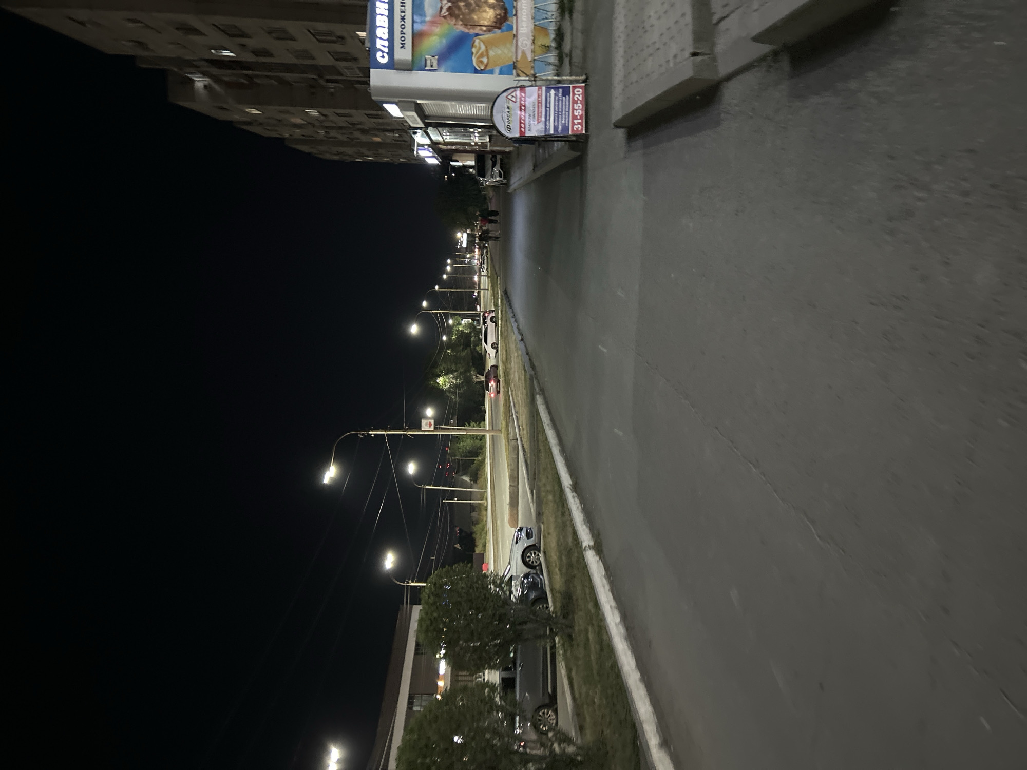 At night, a street in Orsk, Russia, is illuminated by streetlights. A multi-story apartment building with illuminated windows stands on the right side of the street. A store with a sign reading "СЛАВЯНКА" (Slavyanka) and advertising ice cream is visible in the foreground. An A-frame sign with Russian text and a phone number "31-55-20" is placed on the sidewalk in front of the store. Several cars are parked along the street, and a few people are walking in the distance. The street is paved with asphalt, and there are sidewalks on both sides. Trees and bushes line the left side of the street.