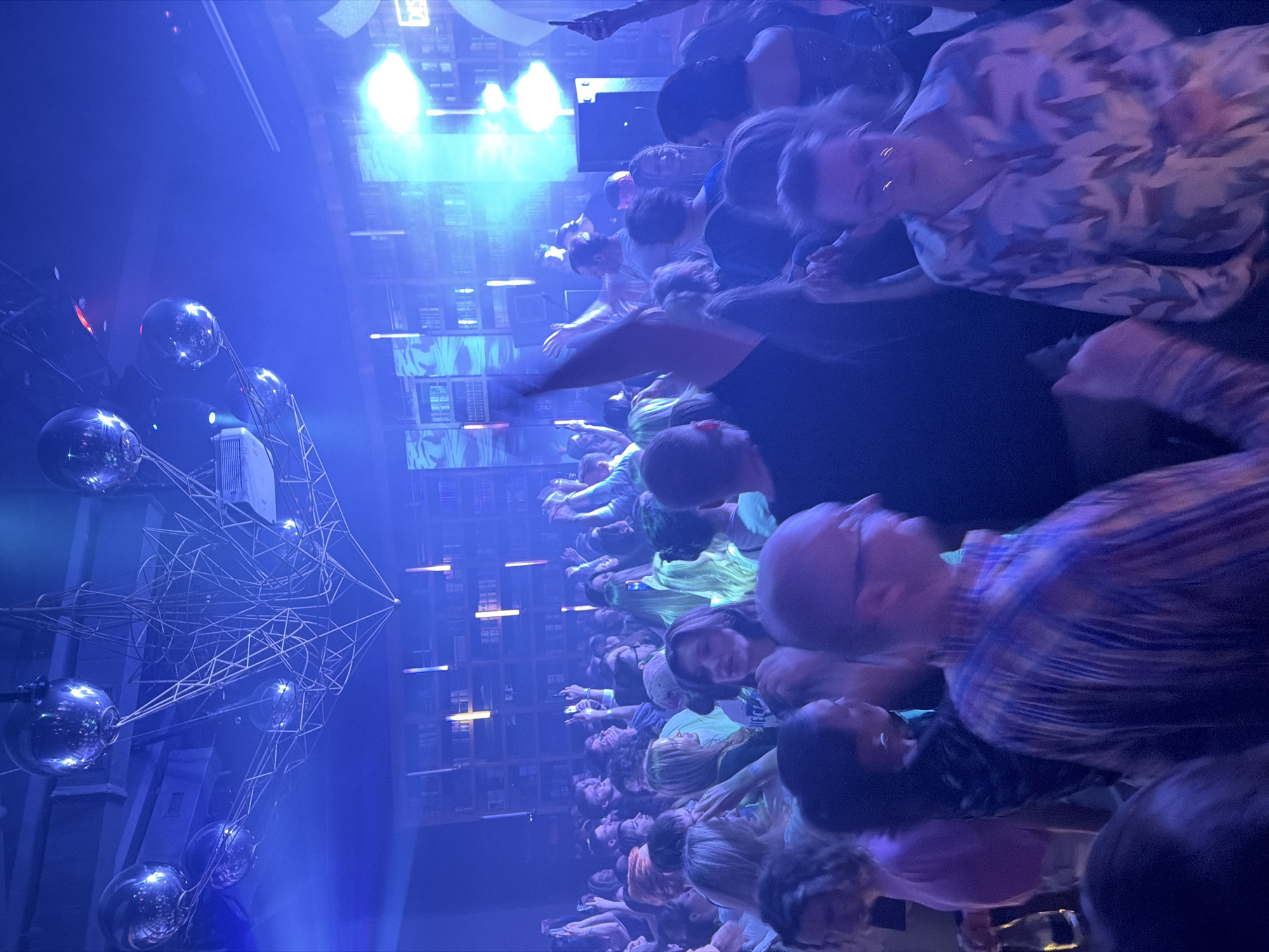 The image depicts a crowd of people in a dimly lit, blue-toned environment, possibly a nightclub or concert venue, suggested by the presence of disco balls and stage lighting. The crowd appears to be looking up at something, possibly a performance or a stage. The location is suggested to be Moscow, Russia.