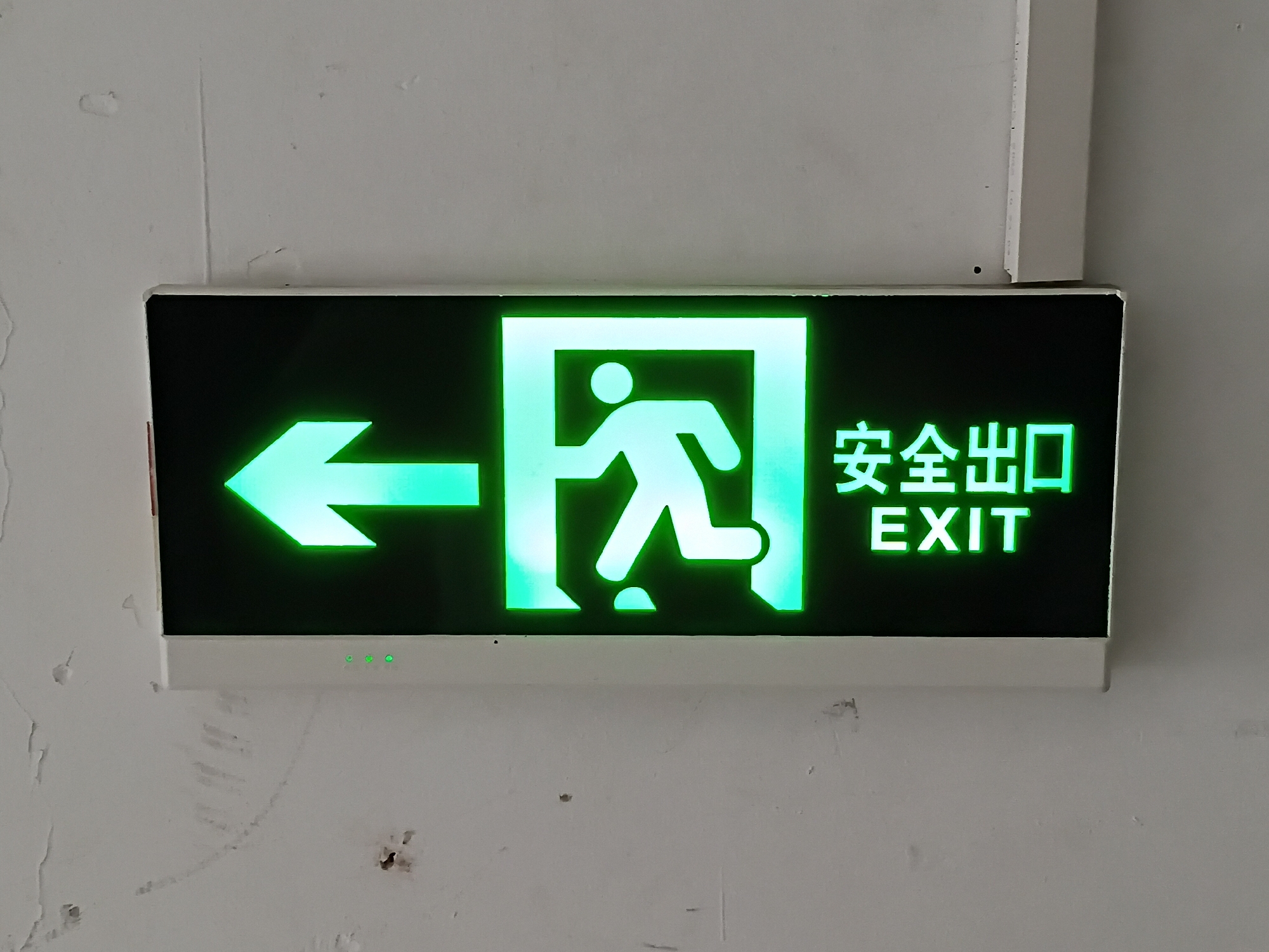 A green illuminated emergency exit sign is prominently displayed on a light-colored, possibly white or light gray, interior wall. The rectangular sign features a bright green pictogram of a person running through an open doorway, with a bold green arrow pointing to the left, indicating the direction of the exit. To the right of the pictogram, the Chinese characters "安全出口" (Ānquán Chūkǒu), which translate to "Safety Exit" or "Emergency Exit," are displayed in green above the English word "EXIT," also in green.
The wall shows signs of minor wear and tear, including subtle scuffs, small marks, and some textural imperfections, suggesting a functional, perhaps industrial or public, indoor environment. The sign itself appears to be an electronic fixture, with small, round, green indicator lights visible along its bottom edge, confirming it is active and powered. The bright green illumination of the sign contrasts sharply with its dark background and the lighter wall, making it highly visible.
The scene depicts a typical safety feature found in public or commercial buildings, providing clear guidance for evacuation. The image focuses directly on the sign, with the wall filling the rest of the frame, hinting at an indoor setting in Shunde District, China. No individuals or specific activities beyond the sign's function are visible. The time of day is not apparent, but the illuminated sign ensures its purpose is clear irrespective of ambient light conditions.