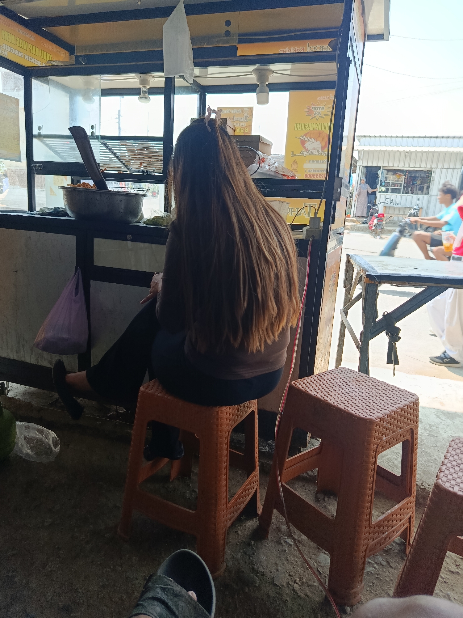 In Kedungmalang, Indonesia, a medium shot captures a person with long, brown hair seated on an orange plastic stool, facing away from the camera, at an outdoor food stall. The person is wearing a dark-colored shirt and dark pants. To the left of the person, a purple plastic bag hangs from the stall's structure. A large metal bowl containing food and a wooden utensil is visible on a shelf in the stall. The stall itself appears to be made of metal and glass, with various signs and advertisements displayed. In the background, a street scene unfolds with a person on a motorcycle and other figures in the distance, suggesting a bustling urban environment. The ground is rough and appears to be unpaved.