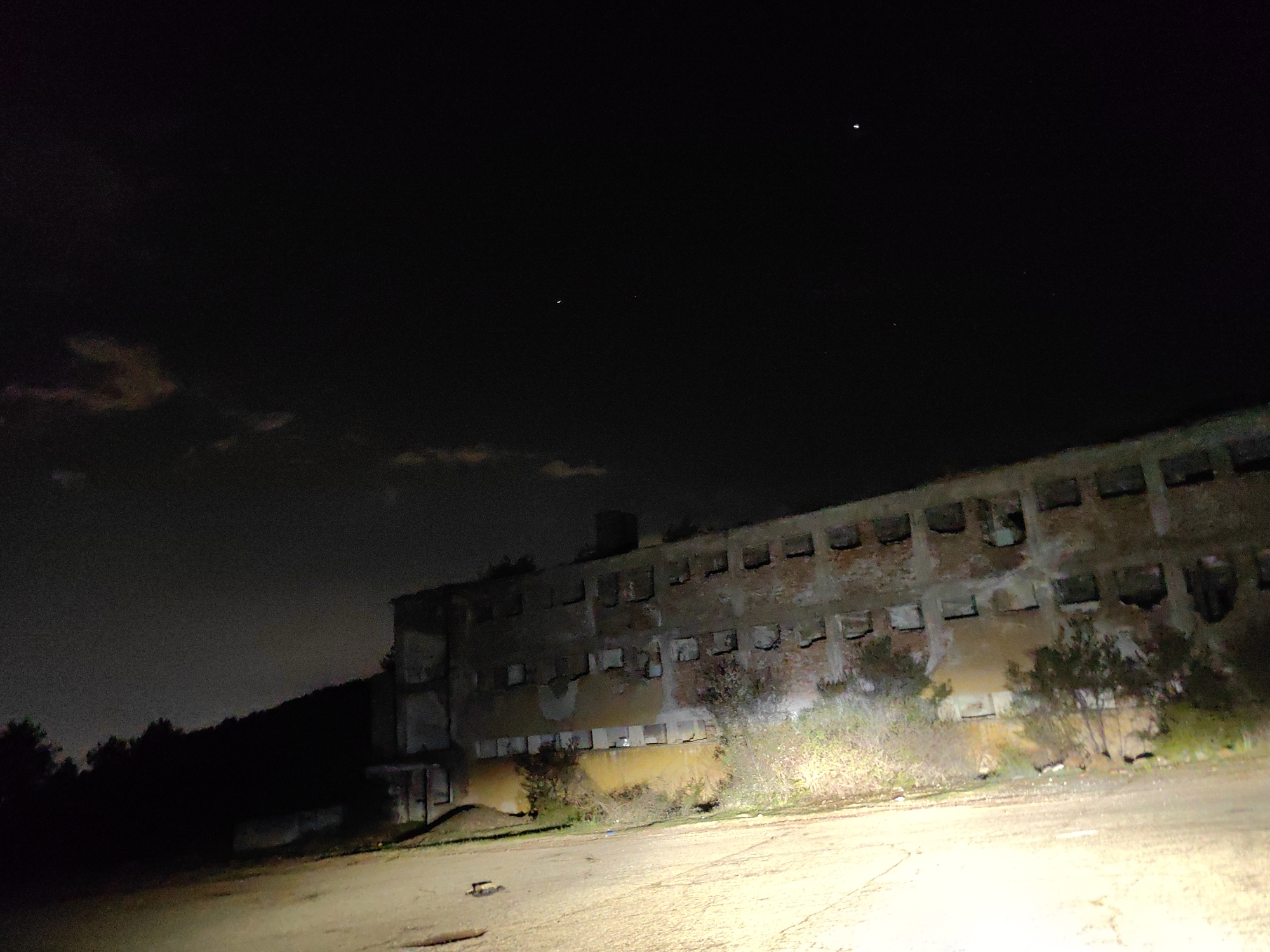 A low-angle, nighttime photograph shows an abandoned brick building with multiple window openings. The building has three floors and appears to be in a state of disrepair, with exposed brick and damaged walls. In front of the building is a dirt or gravel area, illuminated by a bright light source from the bottom of the frame, likely headlights from a vehicle. There are bushes and small trees growing in front of the building. The sky is dark, with a few faint stars visible. On the left side of the image, there is a tree-covered hill. The image is grainy, indicating a low-light photography setting. It is probable that the picture was taken in Sofia, Bulgaria.