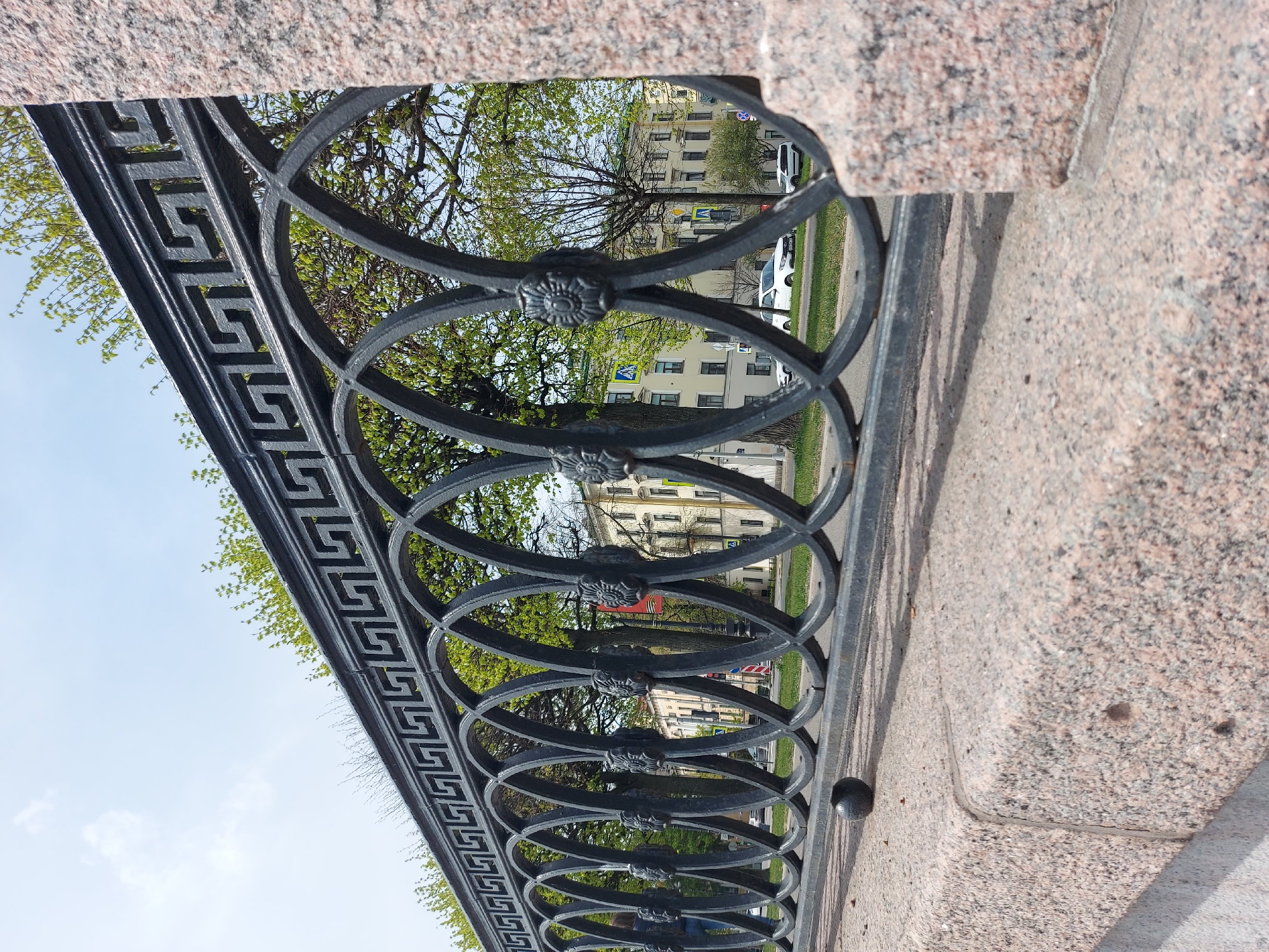 A low-angle, medium shot depicts a cast-iron fence with a Greek key pattern along the top, set against a pink granite embankment, likely along a canal or river in Saint Petersburg, Russia. The fence features a series of overlapping circles, each adorned with a rosette at the point of intersection. Through the fence, one can see a tree with budding green leaves, a building with multiple windows, a crosswalk, cars, and a sidewalk. The sky is visible in the upper left corner.