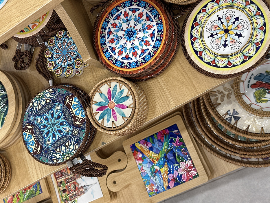 The image displays a retail store shelf in Phú Quốc, Vietnam, showcasing various handcrafted souvenirs and home decor items. The dominant features are multiple circular coasters and trivets, primarily made of ceramic or tile with woven rope or rattan borders, arranged on a light-colored wooden display.
**Objects and Details:**
* **Top Left:** A stack of large circular trivets or hot pads with a prominent blue, white, and brown mandala-like pattern, edged with thick brown braided rope.
* **Below this:** A smaller wooden cutting board features a square tile insert depicting a vibrant, stained-glass style hummingbird with outstretched wings, surrounded by blue, pink, and purple flowers.
* **Center:** A round woven wicker basket contains a set of smaller, lighter-toned coasters, each with a colorful floral design in pink, blue, and orange petals.
* **To the Right of the basket:** A stack of circular trivets, each with a detailed blue, red, and white floral pattern resembling Iznik pottery, edged with a brown rope.
* **Further Right, slightly elevated:** Several flower-shaped coasters with intricate mosaic patterns in blue, yellow, and red tiles are visible.
* **Bottom Right:** A stack of larger circular plates or chargers with rattan-like woven edges. The topmost visible plate features a yellow border and a central blue, pink, and white floral design. Below it, another plate is partially visible with the text "Ao Dai Viet Nam" and illustrations of figures in traditional Vietnamese dress.
* **Background:** Other wooden items and partially visible decorative tiles suggest a broader array of merchandise.
No people are visible. The items are neatly arranged on the wooden shelves, suggesting a static retail display environment.