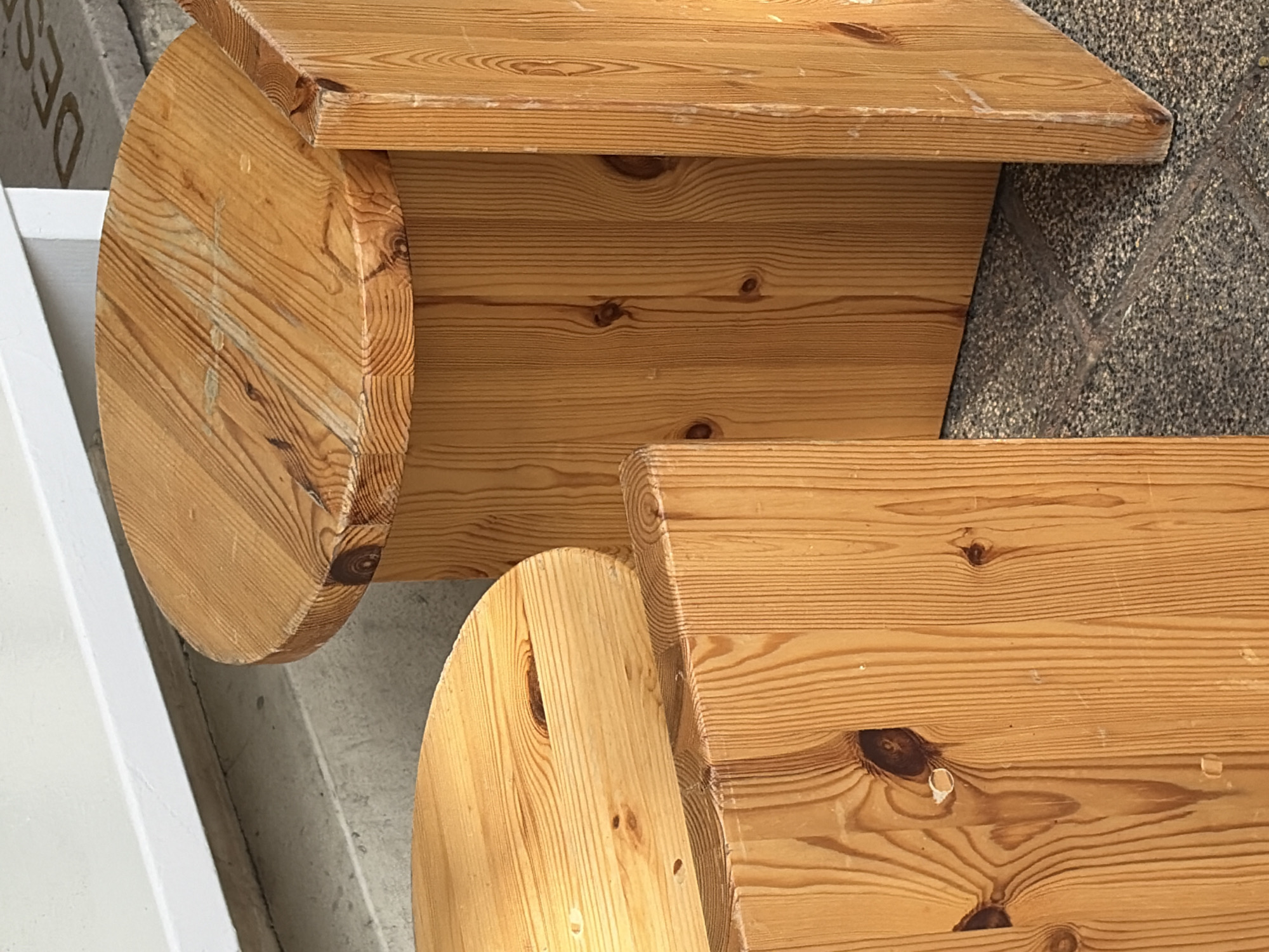 The image shows a close-up of two light-brown wooden tables with a visible wood grain. Both tables have circular support structures. The upper table has a rectangular top, while the visible portion of the lower table suggests a similar design. The setting appears to be an outdoor urban area. No people are visible. There is no information to confirm location in Tel-Aviv, Israel.