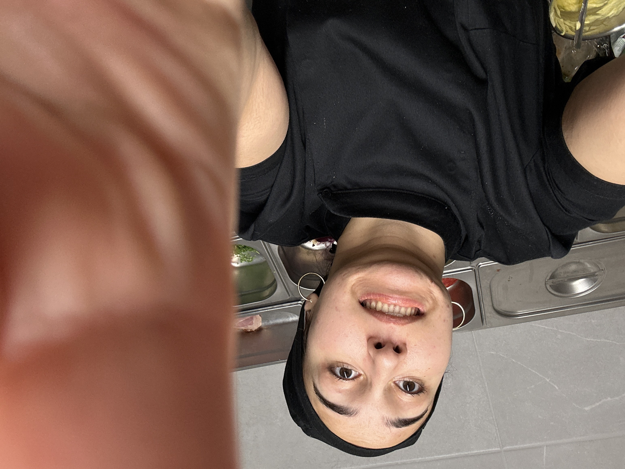 A woman wearing a black head covering and a black shirt is taking a selfie from an upside-down perspective. Her face is visible, and she is smiling. In the background are metal containers and a white tiled surface. The location is likely a commercial kitchen, possibly in the city of Burgas, Bulgaria, though there is nothing definitively confirming the location.