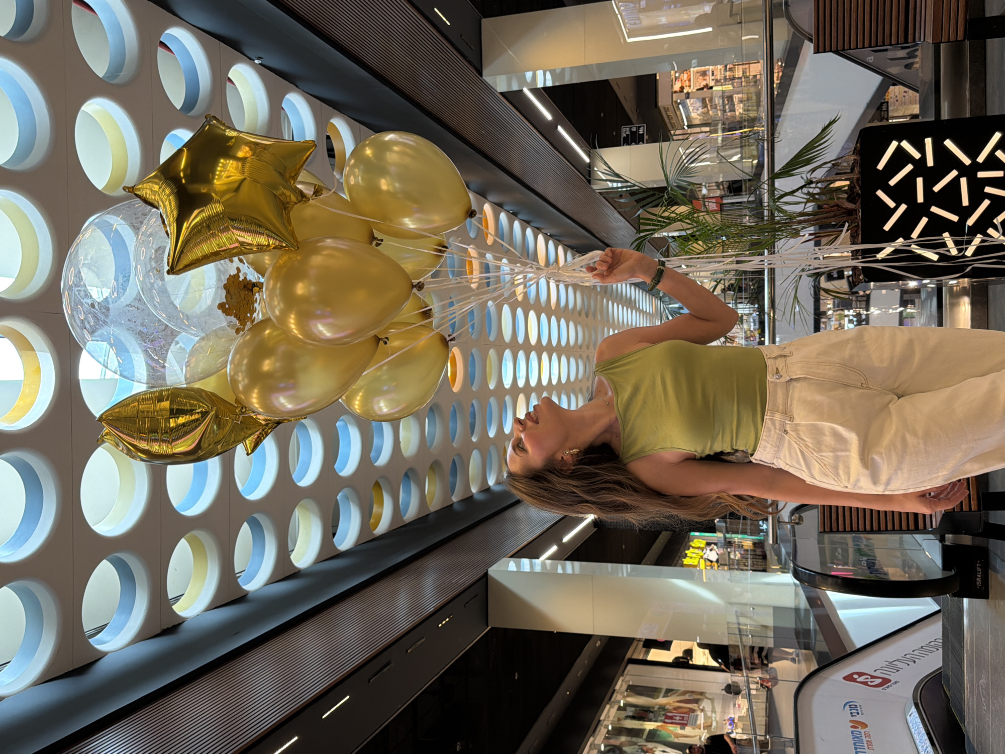 The image depicts a young woman standing in a modern indoor commercial space, likely a shopping mall in Bat Yam, Israel. She holds a large bouquet of balloons, which include several metallic gold round balloons, two clear balloons (one containing gold confetti), and a prominent gold star-shaped foil balloon.
The woman has long light brown hair and is wearing a light green tank top and light-colored jeans. Her left arm is visible as she holds the collection of white balloon strings. Her expression is neutral as she looks towards the left side of the frame.
The background features a distinctive architectural wall composed of a grid of circular openings, some with light blue and light yellow interior accents. To the right, elements of the mall's interior are visible, including escalators, glass railings, a potted plant, and a decorative black panel with illuminated abstract white shapes.
Reflections in glass surfaces indicate other levels or shops within the building. Visible text includes "ISRALIFT" on a dark sign near the escalator, and partial Hebrew text on a white banner on the escalator itself, appearing to include "מכבי" (Maccabi) and "מזרחי" (Mizrahi), consistent with health services or banking advertisements often found in Israeli public spaces. The setting suggests a contemporary urban environment, perhaps celebrating an event or a special occasion with the balloons.