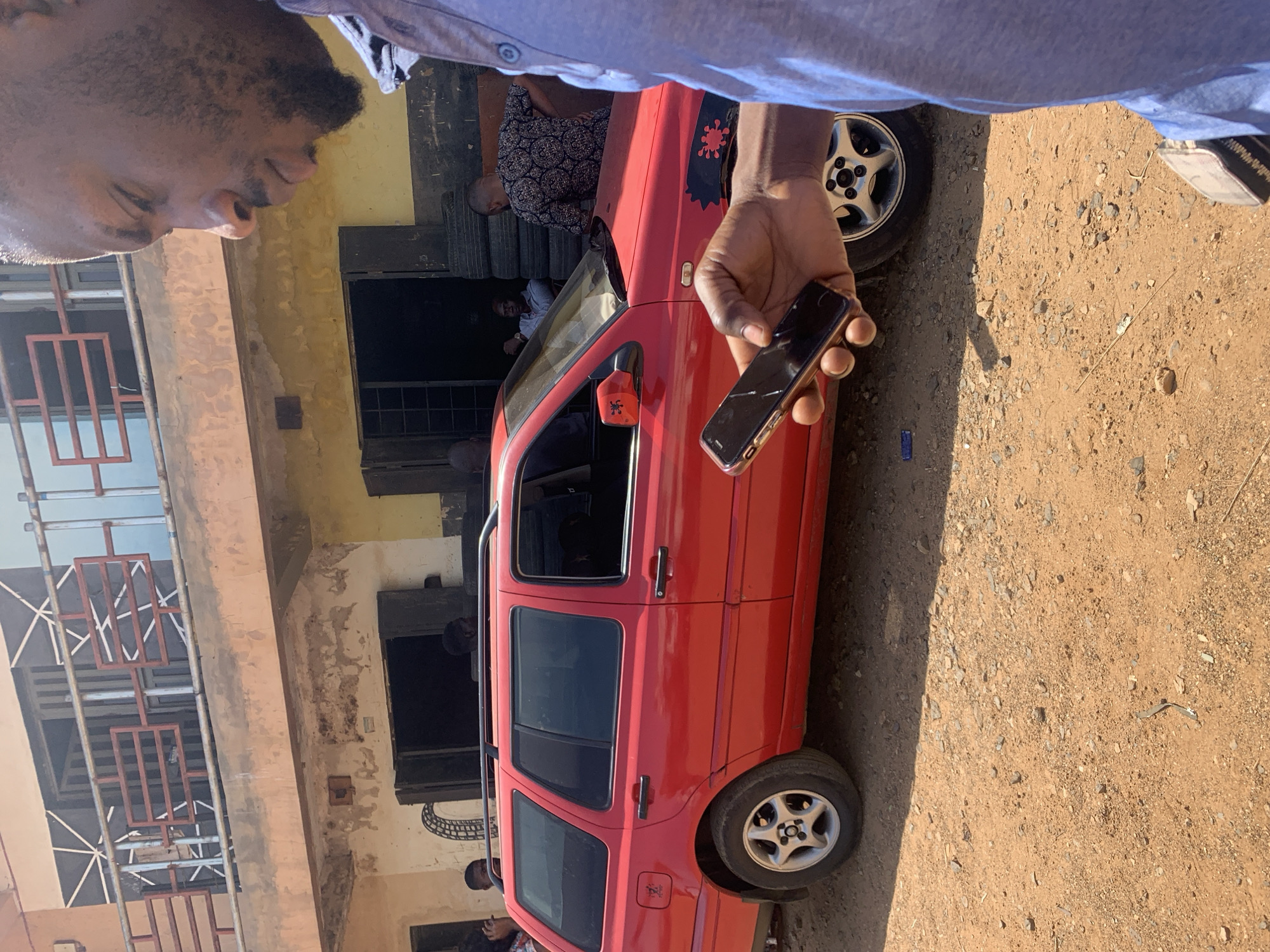 In Rafin Par, Nigeria, the scene depicts a red Volkswagen station wagon parked in front of a weathered, two-story building. A man is visible in the foreground, holding a cracked smartphone in his right hand. The ground is dirt. The building shows signs of age and wear, with peeling paint. Other people are present, one in the car and another in the doorway of the building. Scaffolding is present.