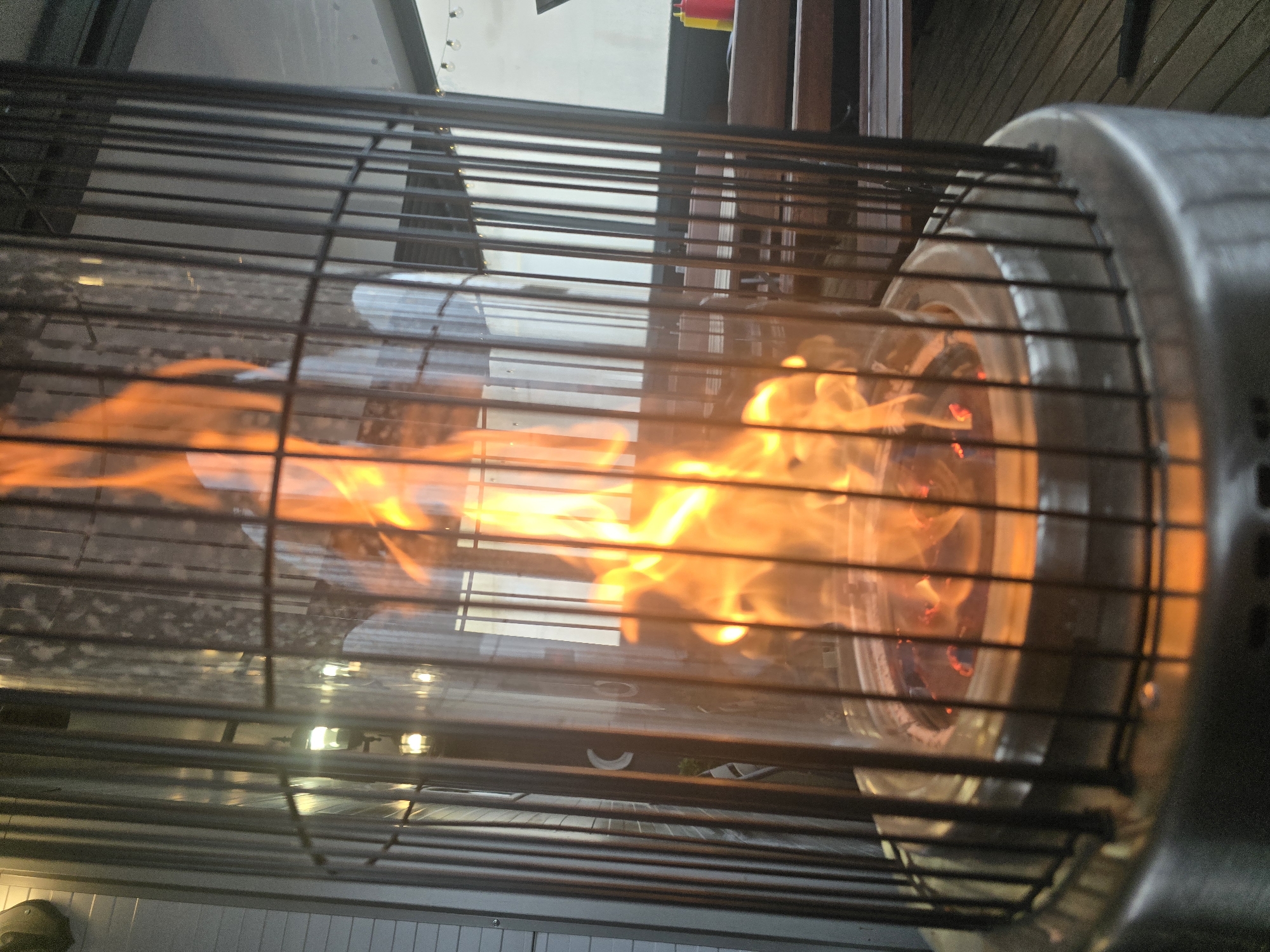 The image shows a close-up of a lit outdoor gas heater. The flames are visible through a metal cage. The heater has a metallic casing around the burner area. The background is blurred, showing an outdoor setting with glimpses of building structures and lights. The location is not identifiable as Samokov, Bulgaria, based on visual information.