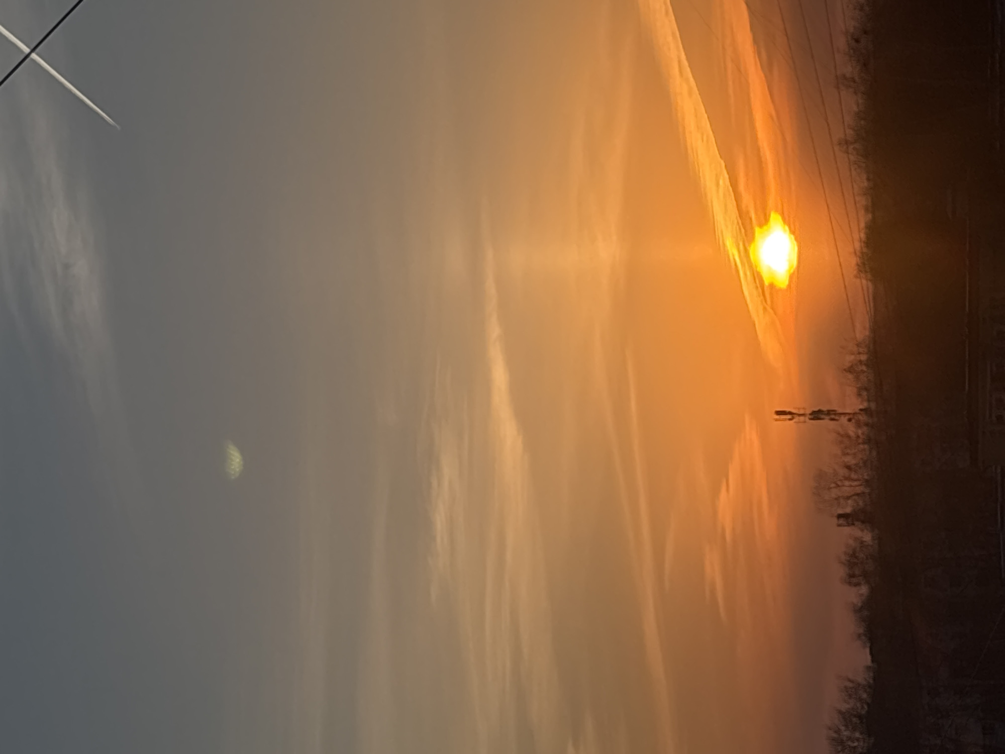 The image depicts a sunset in Chaikovsky, Russia. The sky is a mix of orange and gray, with contrails visible. There is a partially visible moon in the lower left quadrant of the sky. The sun is setting near the horizon, with a bright glow. In the foreground, there are trees and power lines. There appear to be some buildings in the background as well.