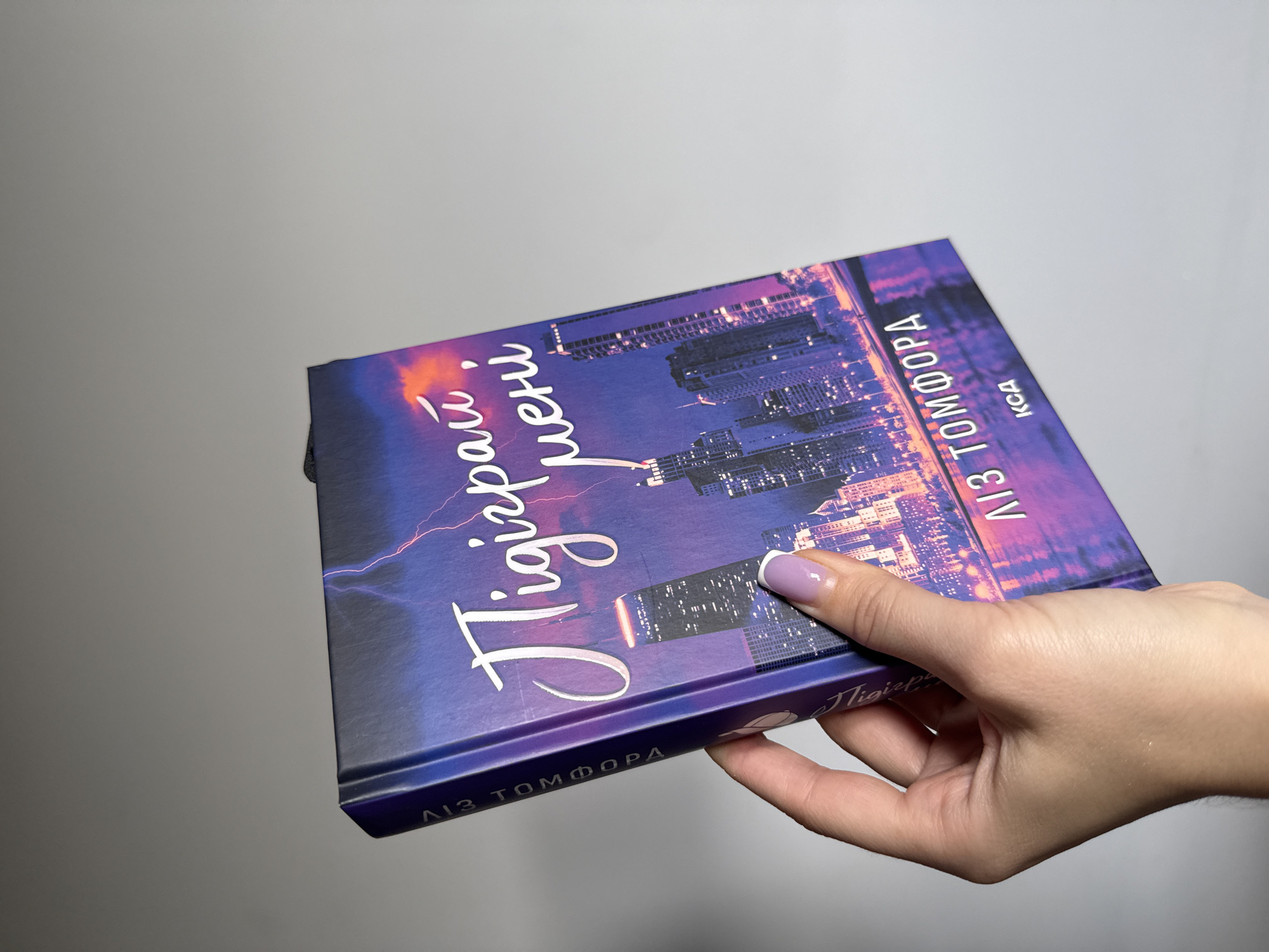 A hand with a light purple manicure holds a book with a dark blue cover against a light gray background. The book's cover depicts a stylized city skyline at night, illuminated by vibrant purple and orange hues and streaks of lightning. The title of the book, written in a flowing white script, appears to be in Ukrainian and reads "Тигрова тіні" which translates to "Tiger's Shadows." Below the title, the author's name is printed vertically: "ЛІЗ ТОМФОРА". A smaller line of text, likely part of the publisher's information or a series name, reads "ксд". The lower edge of the book also displays the name "ЛІЗ ТОМФОРА". The scene is well-lit, suggesting indoor lighting, and the overall impression is one of a modern, slightly dramatic aesthetic for the book cover.
