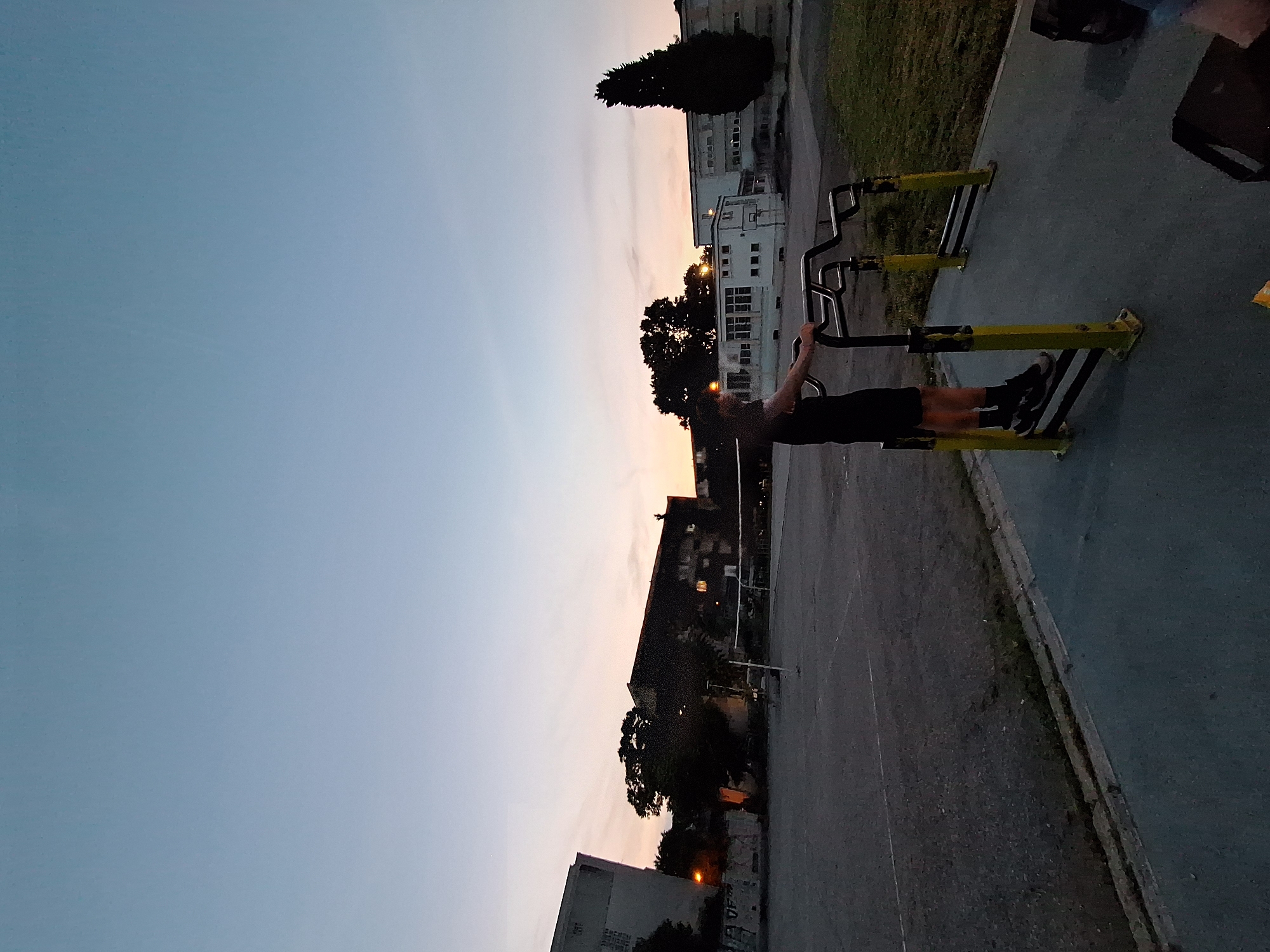 The image captures an outdoor scene at dusk, likely in Varna, Bulgaria, given the context. A lone individual, a person wearing black shorts and a black t-shirt, is standing on an outdoor exercise apparatus composed of yellow posts and black metal bars. The individual is facing away from the camera, with their hands resting on the bars. In the background, a volleyball net is visible on a paved court. Several buildings, including apartment blocks and what appears to be a school or community center, are scattered across the landscape. The sky is a gradient of light blue, transitioning to a soft orange and pink hue near the horizon, indicating the sunset. The overall environment suggests a public space, possibly a park or a schoolyard, designed for recreation.