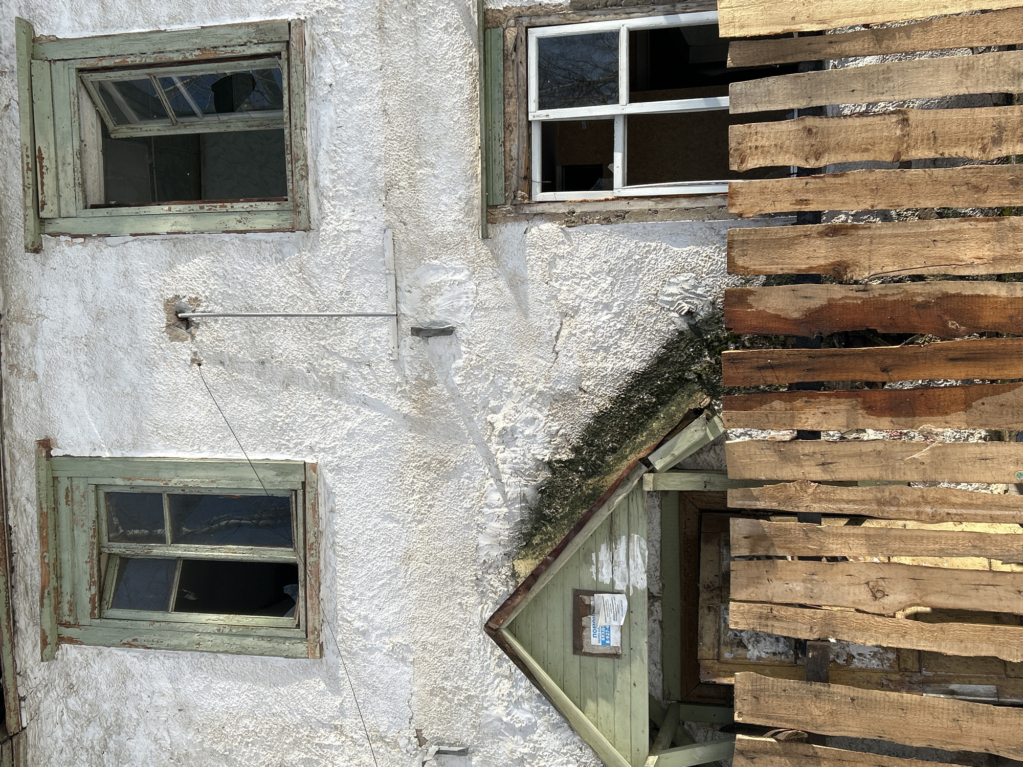An outdoor scene depicts a weathered, off-white stucco or rendered building wall, brightly lit by sunlight, suggesting daytime. The image appears to be rotated 90 degrees clockwise from its natural orientation, showing the wall vertically.
The wall surface is textured and shows signs of age, including cracks, discoloration, and patches of dirt or organic growth. Several window openings are visible, along with some attached wooden structures.
In the upper left quadrant, a rectangular window with a wooden frame painted a faded, peeling light green or teal color is partially open. The top pane is tilted inwards, and several glass panes are broken or missing, revealing a dark interior. Reflections of bare tree branches or sky are visible in the remaining glass.
To the upper right, another window is partially obscured by a series of horizontal wooden planks. This window has a white frame, possibly made of PVC or painted wood, and one of its panes is entirely absent, creating an empty rectangular opening. The frame also appears worn.
Below the upper left window, a thin, horizontal metal rod or pipe extends from a small, rough opening in the wall. A dark wire hangs downwards from it.
In the lower left quadrant, a third window, similar in style and color to the upper left one, is mostly closed. Its faded light green/teal wooden frame also shows signs of deterioration, and a lower right glass pane is broken or missing, showing a dark interior.
On the right side of the image, a stack of rough, weathered wooden planks, resembling pallet wood, are nailed horizontally to the wall. They vary in color from light brown to darker, water-stained hues. These planks cover part of the wall and extend downwards to a gabled wooden structure below.
This lower structure is a small, lean-to or porch roof, also painted in the faded light green/teal color, situated at the base of the main wall. Its sloped roof is dark and heavily covered with moss or algae. On the front face of this structure, a small, rectangular opening or window has a piece of white paper taped inside. Text in Cyrillic script is visible on this paper; the word "покуп" (pokup, likely meaning "buy" or "purchase") is partially discernible.
Overall, the scene portrays a neglected or abandoned building with various elements in disrepair, characterized by peeling paint, broken glass, and weathered materials.