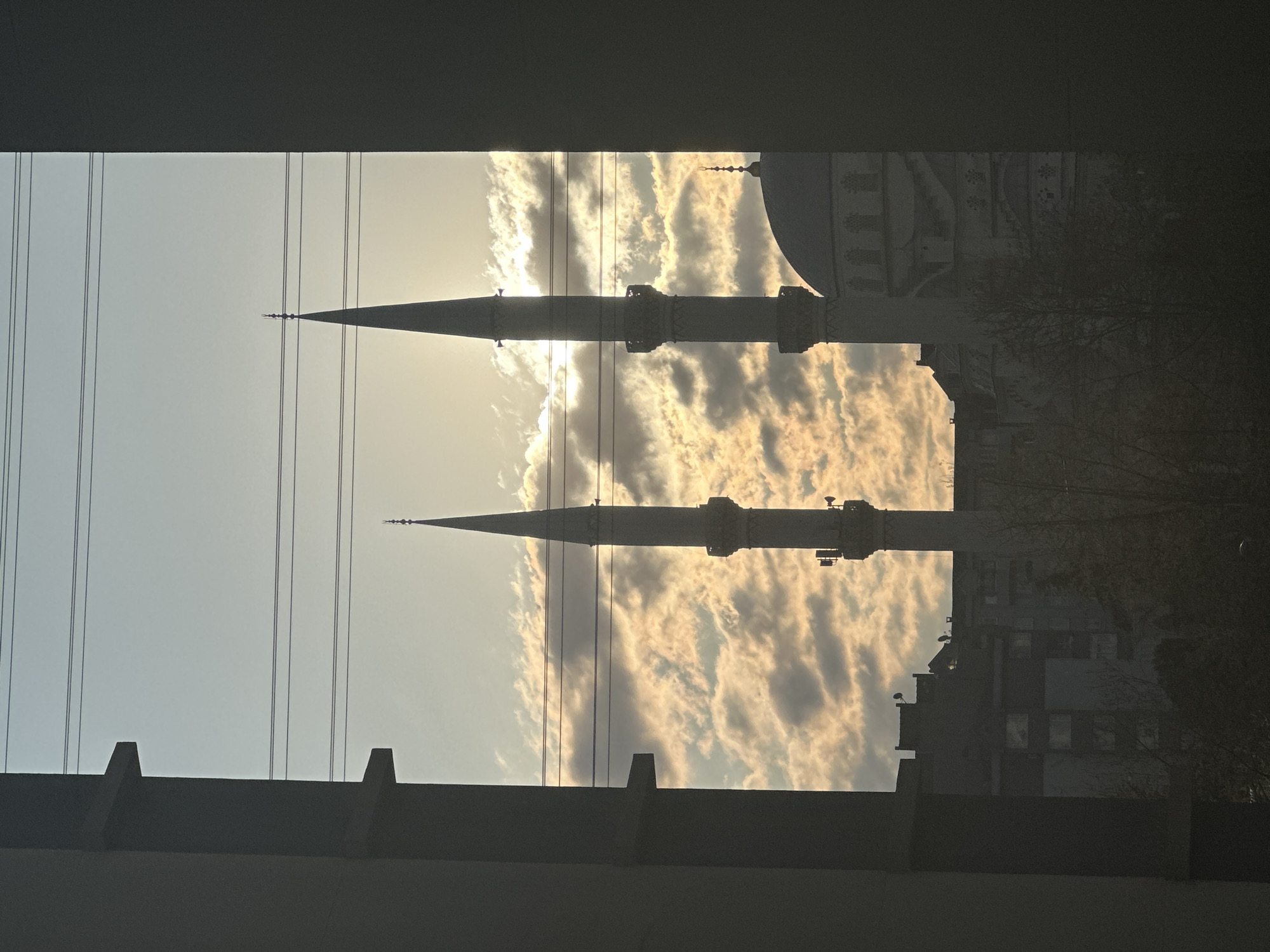 The image shows a view of a mosque with two minarets and a dome, framed by a concrete structure, likely a bridge or building. The sky behind the mosque is filled with clouds, illuminated by the sun. Power lines are visible on the left side of the frame. The mosque is located in Esenyurt, Turkey. Buildings and trees can be seen in the background to the right. The lighting suggests it is either sunrise or sunset.