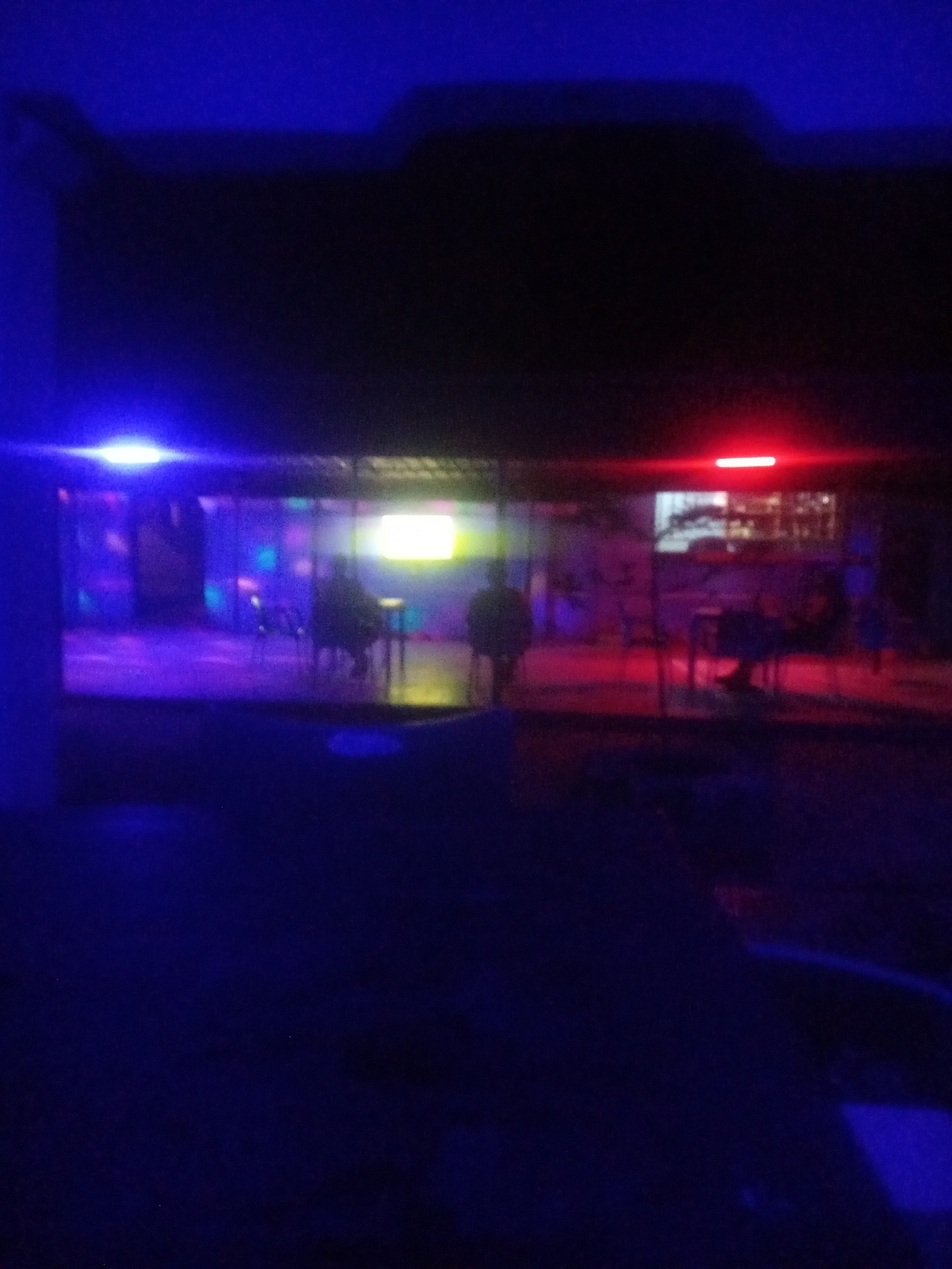 The image depicts an outdoor scene at night in Sabon Gari, Nassarawa, Nigeria. The foreground is dominated by dark blue hues, suggesting a night sky or dimly lit surroundings. In the mid-ground, a brightly lit structure, possibly a bar or restaurant, is visible. The structure features purple and pink lighting, creating a vibrant atmosphere. Several individuals appear to be present inside and around the establishment, though their features are indistinct due to the low light conditions. The lighting includes a prominent blue light on the left and a red light on the right, illuminating the area. The overall impression is that of a lively nighttime social gathering.