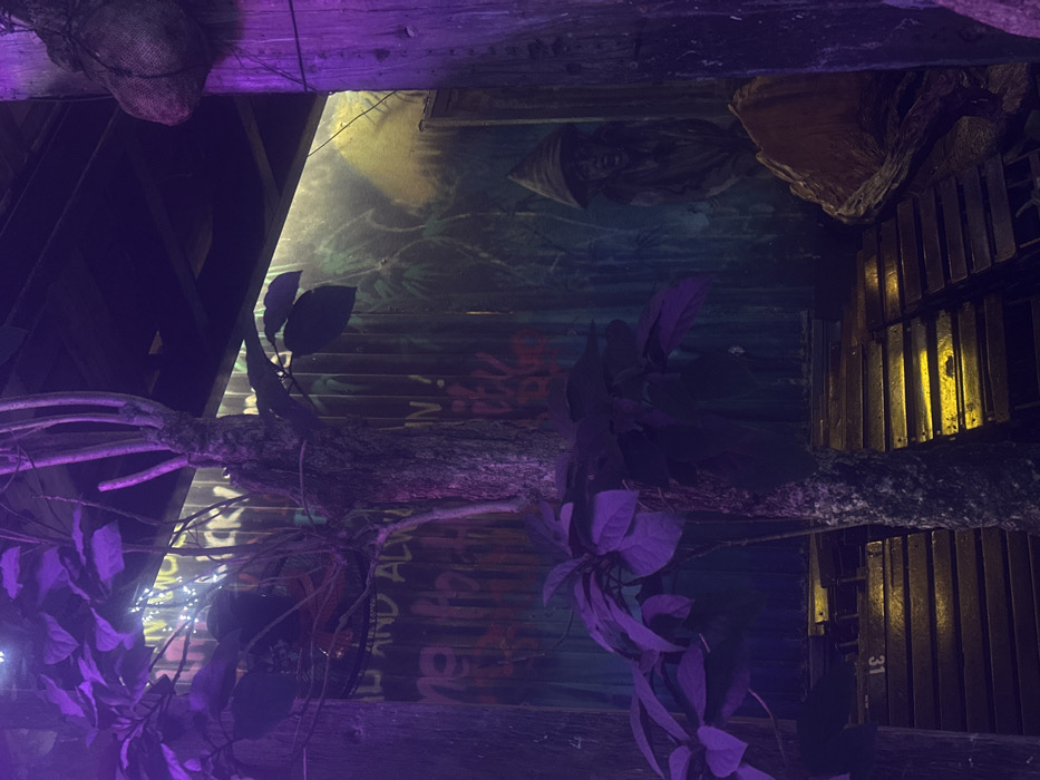 The image depicts an interior or semi-enclosed space, likely a commercial establishment, within Thuận Hóa Ward, Vietnam, illuminated by prominent purple and yellow artificial light.
A central tree with a dark, textured trunk and broad leaves dominates the midground, with its foliage appearing purple under the lighting. A metal fan cage is partially visible through the upper left branches of the tree. The background consists of a corrugated metal wall. The left portion of this wall displays graffiti-style text, including "AND ALWAYS" and "IN!", while the right portion features a painted mural of jungle foliage, with a bright yellow light source illuminating the upper right section. Against the right side of this mural, a dark, sculptural figure of a person wearing a conical hat (nón lá) is visible.
To the far right, a vertical wooden post supports a burlap-like sack suspended near its top. The upper section of the image reveals dark wooden structural elements. At the bottom of the frame, multiple rows of stacked wooden chairs or benches are arranged, strongly illuminated by yellow light. One chair in the foreground displays the number "31". A large piece of natural wood, resembling a log, rests on the ground to the right of the stacked seating. No individuals are present in the scene.