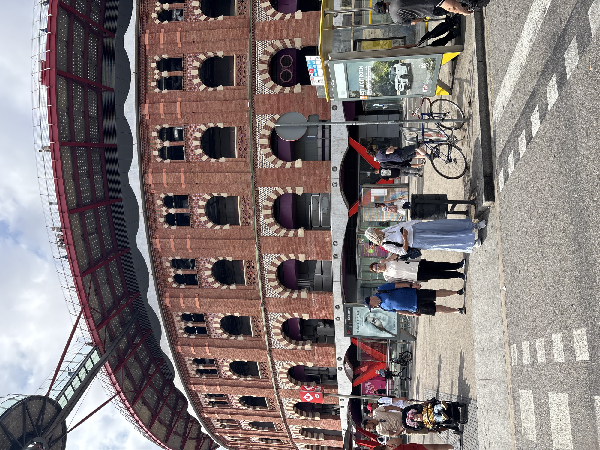 The image captures a street scene in front of the Las Arenas de Barcelona, a former bullring turned shopping center, located on Gran Via de les Corts Catalanes in Barcelona, Spain. The iconic red brick building with its distinctive arched windows dominates the background. A modern metal and glass walkway or bridge extends from the top of the building, showcasing a panoramic view of the city. In the foreground, a group of people are visible. A family with a baby in a stroller stands on the right, near a bus stop. A man in a blue shirt and shorts, a woman in a white top and black pants, and another woman in a light blue skirt and white top are also present. A bicycle is parked next to a bus stop shelter displaying an advertisement for Citroen. Pedestrians are crossing a marked crosswalk, with the urban environment of Barcelona evident through the road and pavement. The sky is partly cloudy.