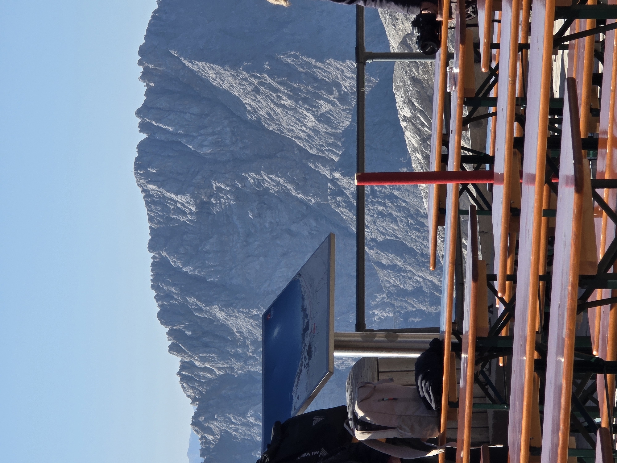 The image depicts an outdoor seating area with several picnic tables in the foreground, set against a backdrop of a large, rocky mountain range under a clear blue sky. The tables, with orange painted wooden tops and green metal legs, appear to be in a restaurant or cafe setting. In the middle ground, a person is partially visible on the right, holding a camera. A red pole stands vertically between the tables and a metal railing. To the left, a signpost with a reflective blue surface is mounted on a metal pole, and a backpack rests on a table. The mountain landscape is rugged and imposing, suggesting a high-altitude location. This scene is likely from the Zugspitze, Germany's highest peak, located near Garmisch-Partenkirchen.