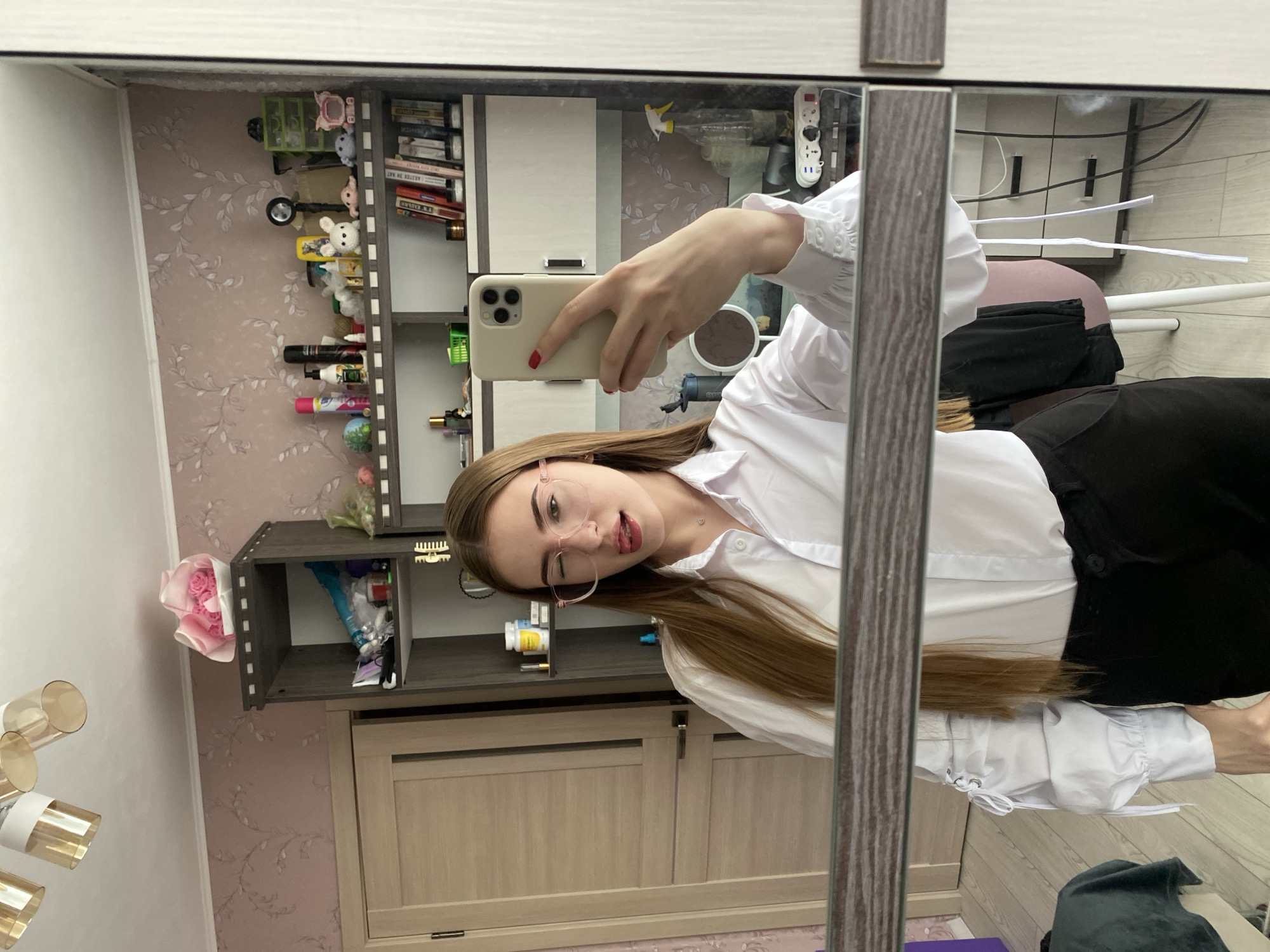 A young woman with long, brown hair and glasses is taking a mirror selfie in a room. She is wearing a white collared shirt with ruffled sleeves and black pants. Her tongue is sticking out, and she is winking. The room appears to be a bedroom with a shelf unit filled with various items, including books, toiletries, and decorative objects. There is a door and a patterned wallpaper visible in the background. The lighting suggests it is daytime. The location is stated as Дьяково, Russia.
