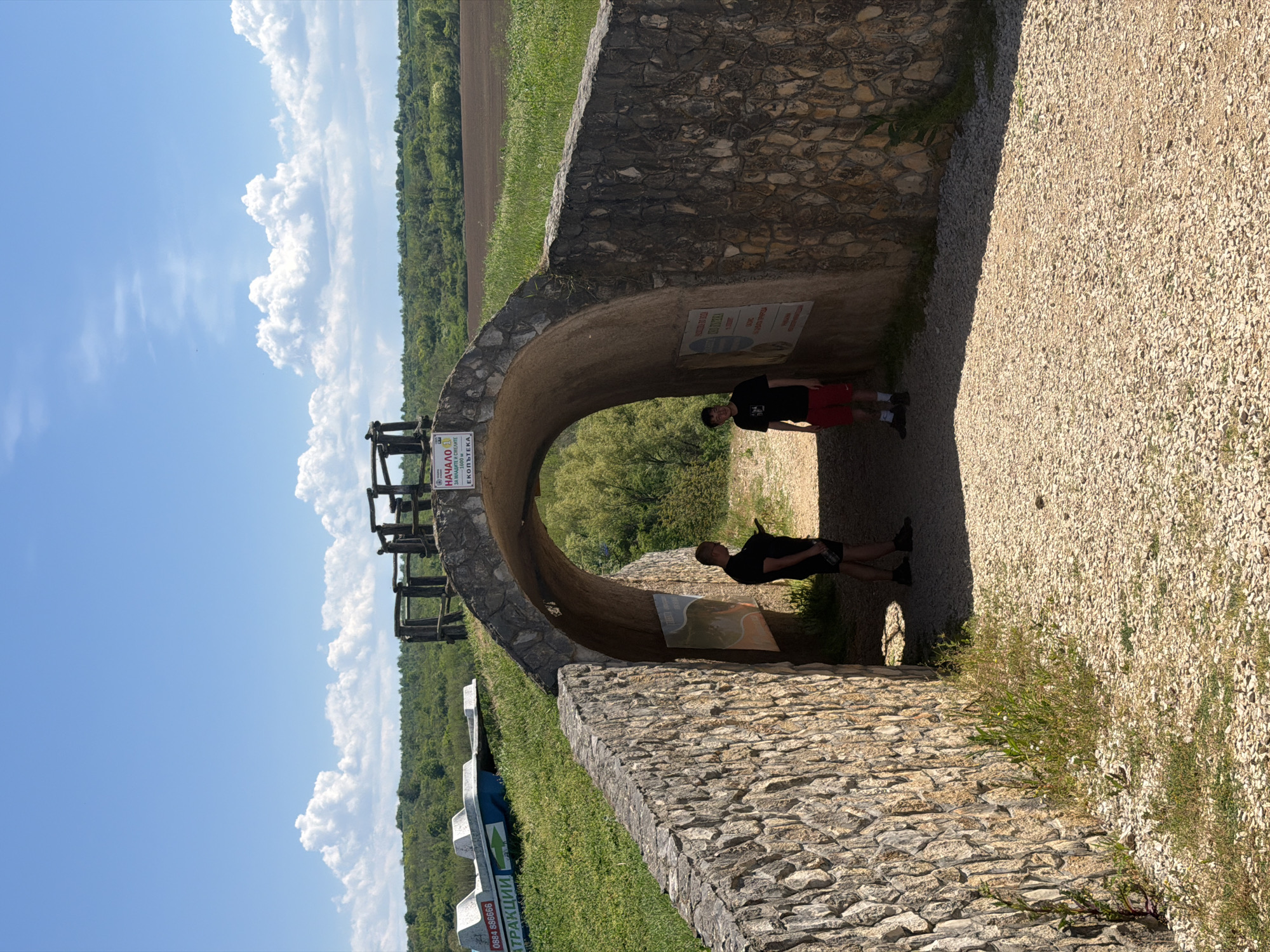 The image shows a stone tunnel-like structure located in Lukovit, Bulgaria. Two people are standing inside the tunnel. One person is wearing a black t-shirt and red shorts, and the other is wearing a black t-shirt and black shorts. There are informational signs on the walls of the tunnel. Outside the tunnel, there is greenery and a view of fields and trees under a blue sky with white clouds. To the left of the tunnel entrance is an observation structure, and what appears to be a guide arrow sign. The ground near the tunnel is covered in gravel.