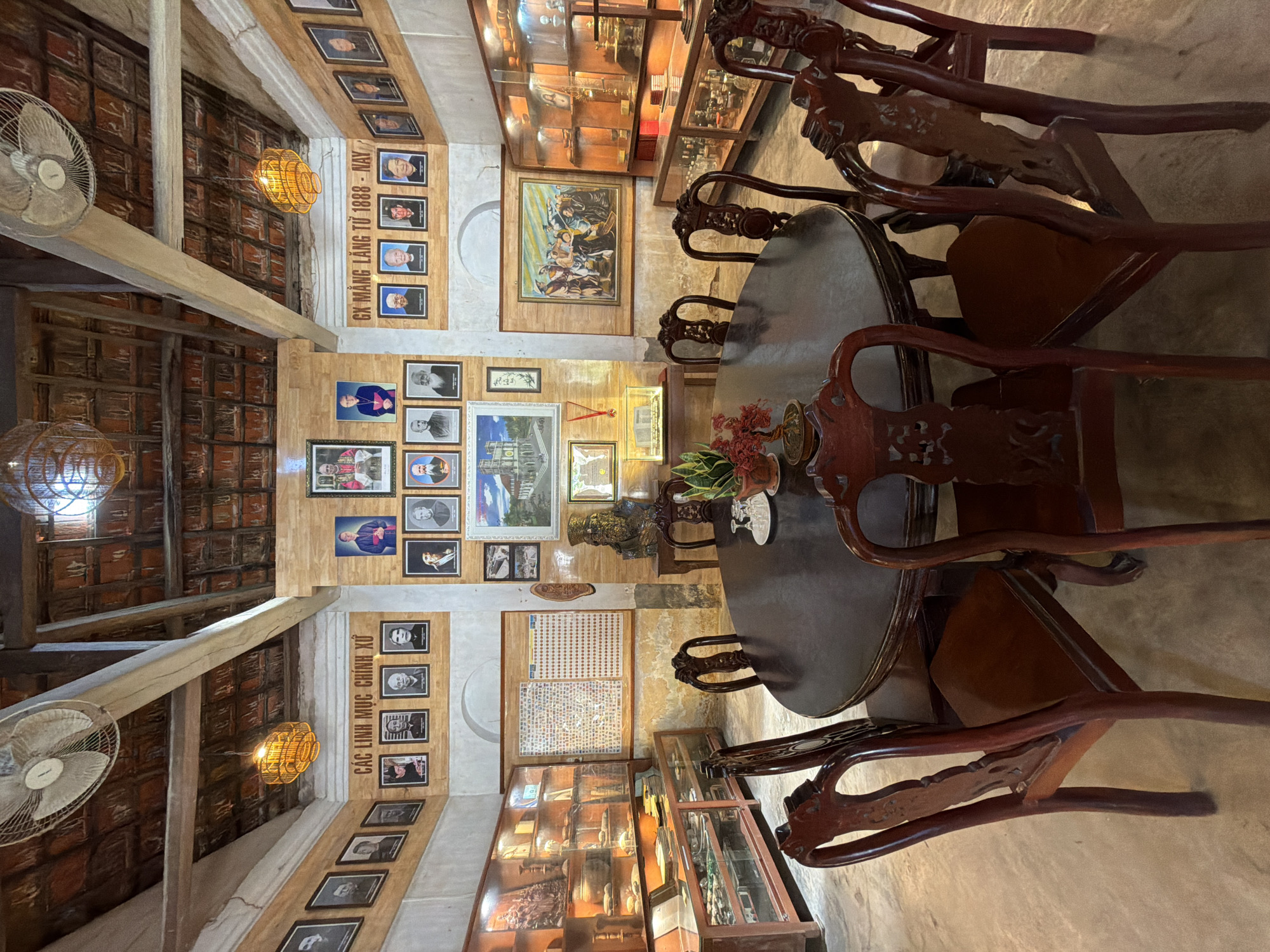 The image shows an interior scene of what appears to be a museum or historical exhibit room in Hội Tín, Vietnam. A large, dark, oval wooden table with eight ornately carved wooden chairs surrounds it. On the table, there are a few objects, including a small plant and two glasses.
The walls of the room are lined with framed photographs and paintings. On the left wall, there are display cases filled with artifacts, including statues and what appear to be religious items. The ceiling is made of wooden beams and terracotta tiles, with two ceiling fans and woven bamboo light fixtures.
On the back wall, there are multiple rows of framed portraits, many of which appear to be of religious figures. There are also two large paintings on the walls, one depicting a group of people and another of a building. In the center of the back wall, there is a large framed display with a grid of small images.
The overall impression is of a well-preserved historical space, likely dedicated to showcasing the history and figures associated with the Hội Tín area. The lighting is warm, contributing to a sense of history and reverence.