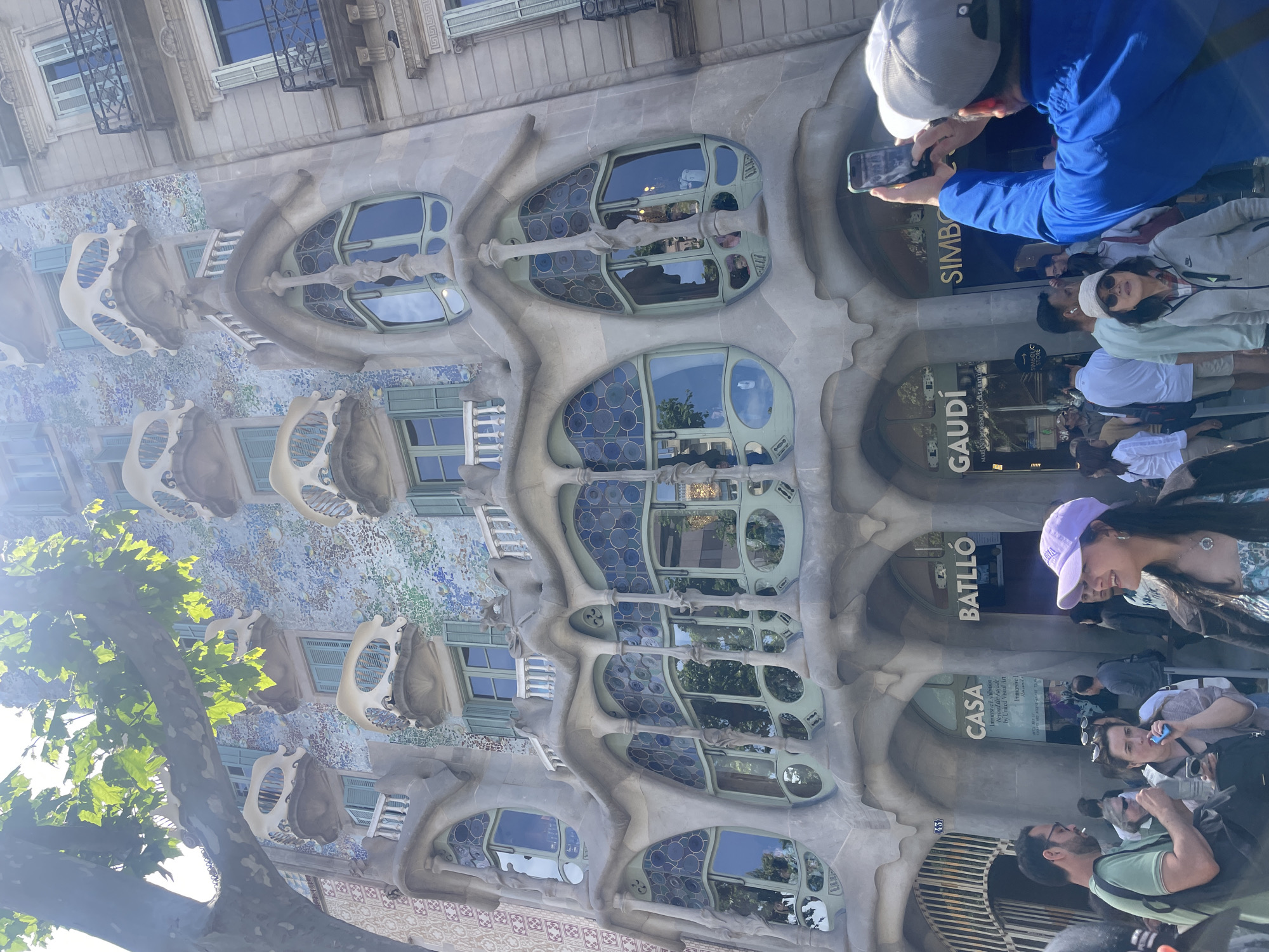 This is a photograph of Casa Batlló, a famous building in Barcelona, Spain, designed by Antoni Gaudí. The façade of the building features undulating lines, oval windows, and skeletal-like balconies. The exterior is decorated with mosaic tiles in shades of blue and green.
In the foreground, there is a group of tourists and locals who are standing in front of the building, observing it. One person is taking a photo of the building with their phone. The entrance of the building displays the name “CASA BATLLÓ” vertically, above the words “GAUDÍ.” The signage next to the entrance indicates “SIMBOLIC STORE”. Trees on the left side of the image partially obstruct the view of the building.