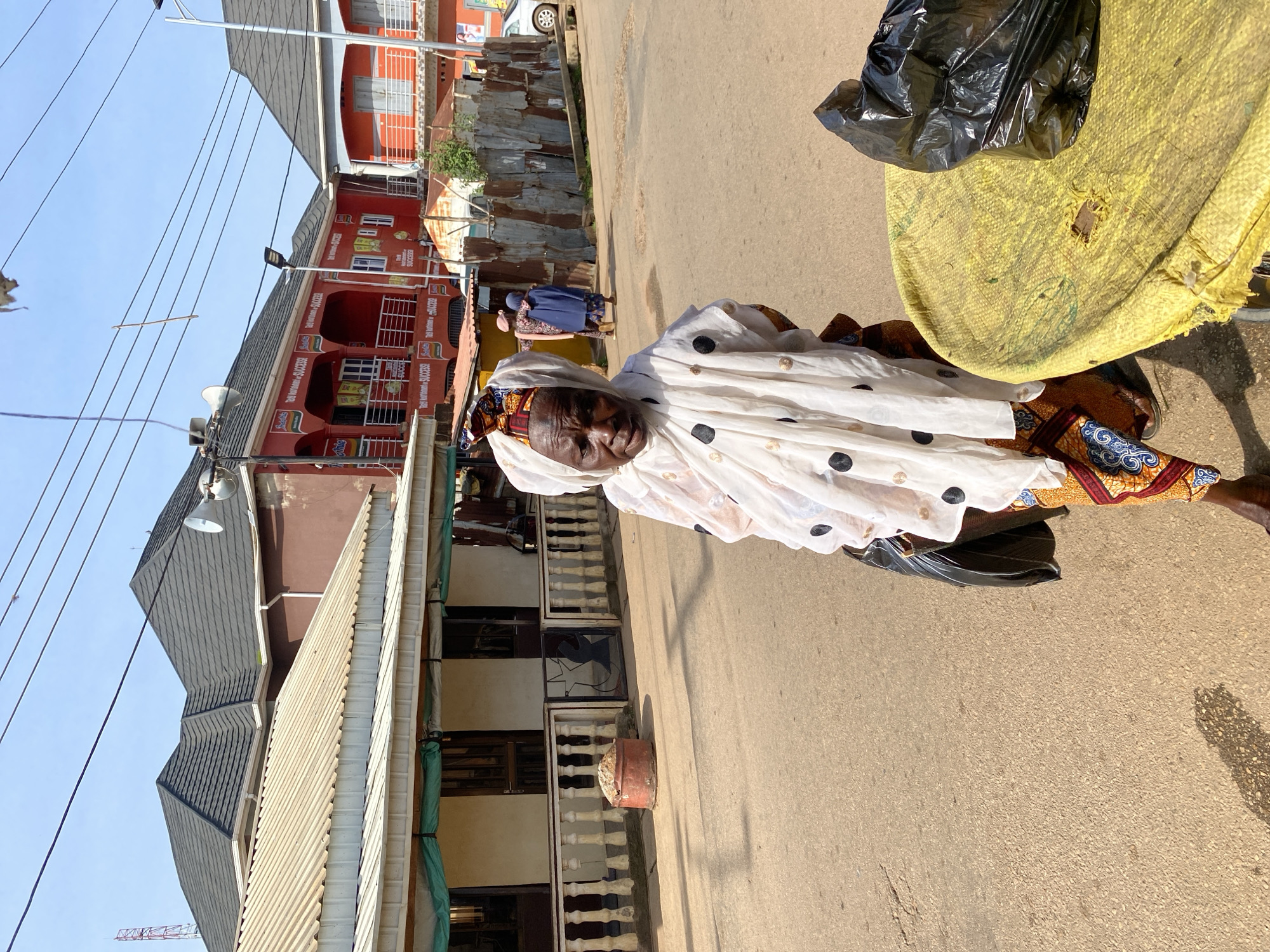 An older Black woman, wearing a white headscarf with black and gold polka dots and an orange and blue patterned skirt, stands in the foreground of a street scene in Jos, Nigeria. She is holding a black plastic bag in her left hand. To her right, a round yellow table covered with a rough textured material has a black plastic bag resting on it. The street is paved with asphalt, with some cracks and debris visible. In the background, there are several multi-story buildings with red and maroon facades, featuring balconies and signs with text. One sign appears to be for "Indomie Success." Further in the background, there are corrugated metal fences, a white car, a street lamp, and power lines. The sky is clear and blue. The overall impression is a bustling urban environment.