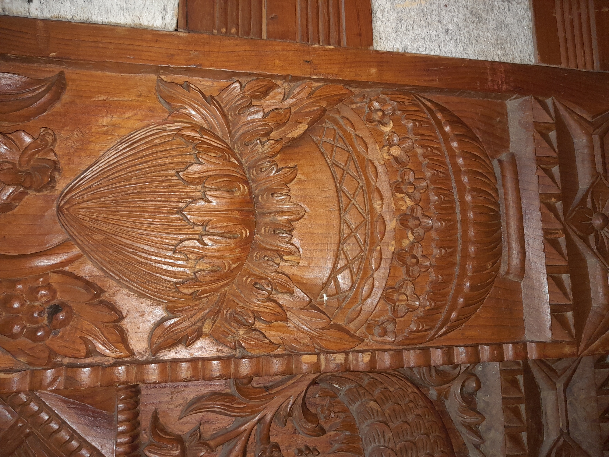 The image displays a close-up, detailed view of intricate wood carving on a piece of furniture or architectural element. The carving features a central motif resembling a lotus flower emerging from a decorative vase. The lotus petals are finely detailed with striated lines, and the vase itself is adorned with geometric patterns and smaller floral designs. Flanking the central motif are stylized leaf and floral elements. The wood appears to be dark and polished, showing some signs of age and wear. The background suggests a rough, textured surface, possibly stone or plaster. The scene is lit to highlight the carvings. No people or specific location details are discernible.