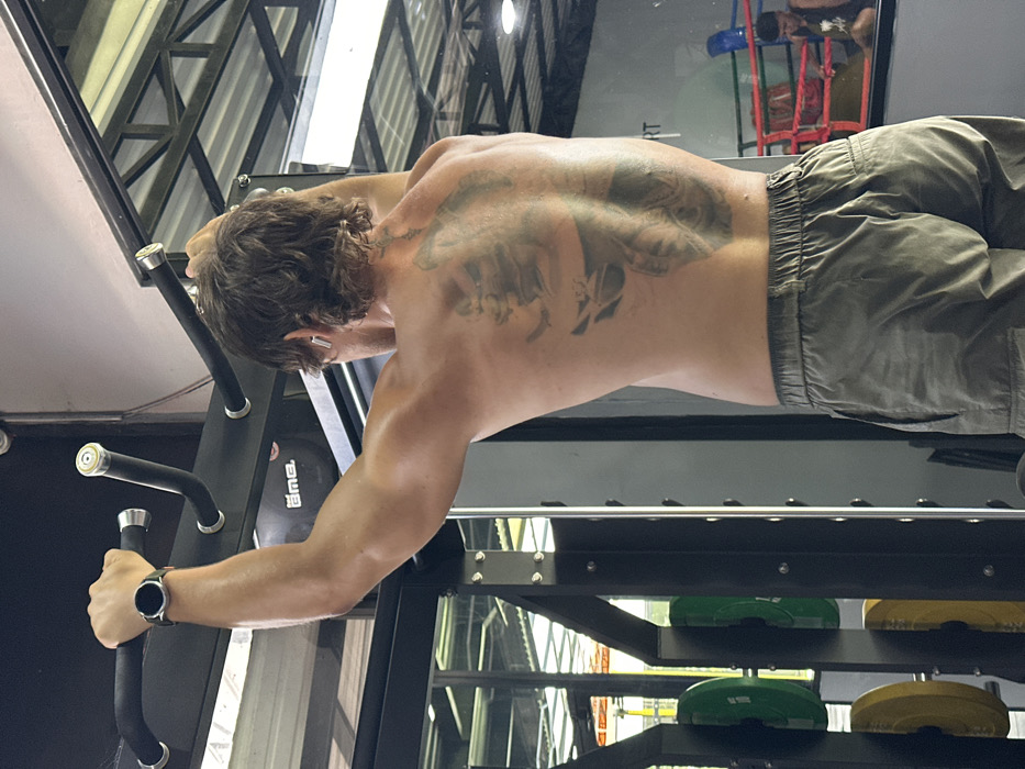 A male individual is depicted from the back, shirtless, performing an exercise on a black multi-grip pull-up bar within a commercial gym setting in Tibubeneng, Indonesia. The individual, exhibiting a muscular physique and light-brown, wavy hair, grasps the bar with an overhand grip, arms extended. He wears olive green cargo-style shorts, a black smartwatch with a silver bezel on his left wrist, and white wireless earbuds in both ears. A large, dark tattoo featuring skeletal and possibly other imagery covers his upper and mid-back. To the left, green and yellow weight plates are stacked on a black metal storage rack. In the background, a large mirror reflects a boxing ring with blue ropes and a red corner post, within which a second male individual is partially visible, seated or squatting. The gym features a corrugated metal ceiling and an overhead black object marked "ama".