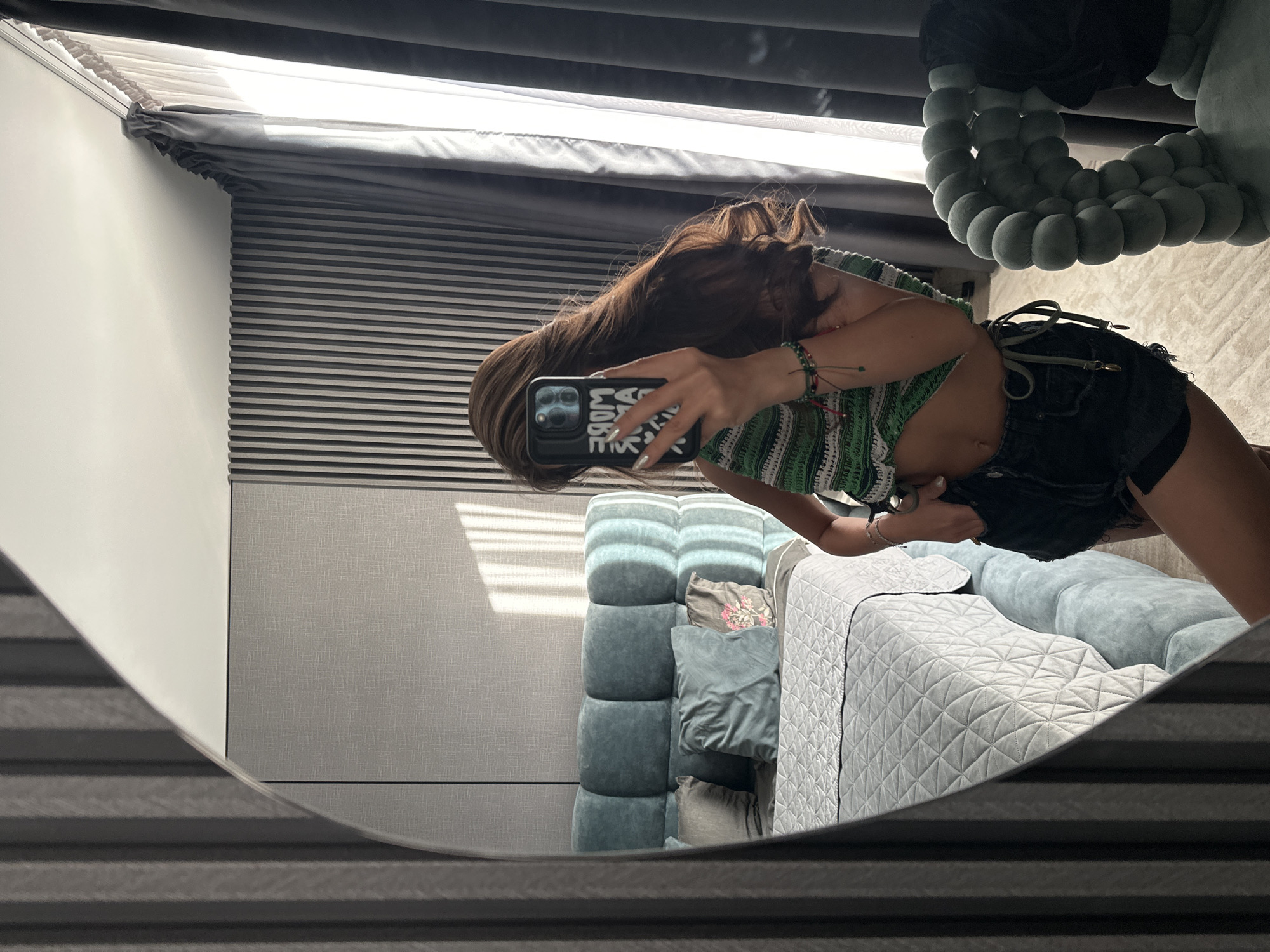 A person is taking a mirror selfie in a bedroom. The person has long brown hair and is wearing a green and white striped crochet top and black denim shorts. They are holding a smartphone with a case that has white lettering. In the background, a bed with a light blue tufted headboard and a quilted grey duvet is visible. There are also pillows on the bed. To the right, a grey upholstered chair with a rounded, bubbly design is present. The room has modern wall paneling and dark grey curtains. The image appears to be taken indoors, likely in a residential setting. There is no discernible information indicating the location is Sofia, Bulgaria.