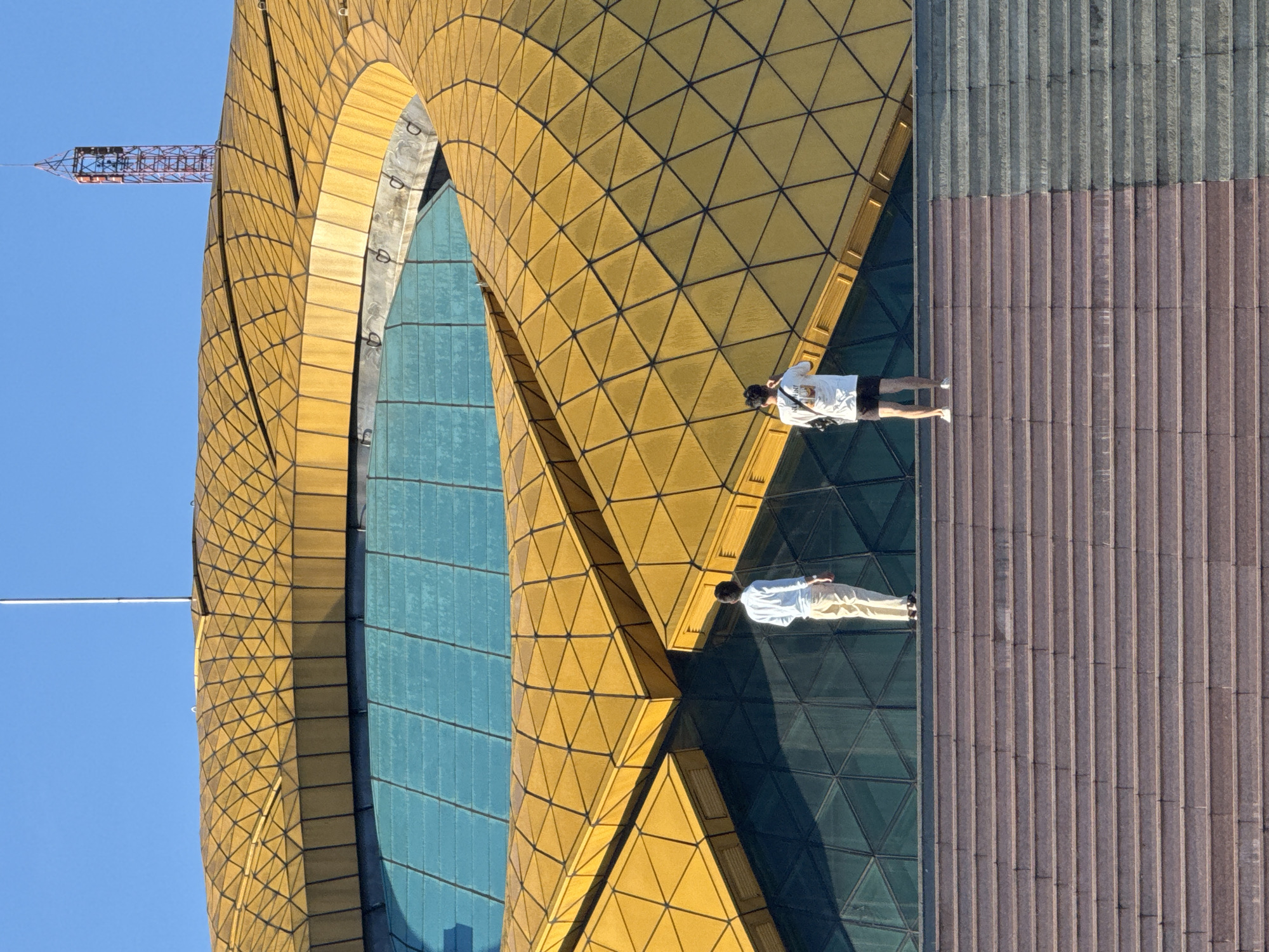 The image shows the exterior of the Da Lat Palace Convention Center, located in Da Lat, Vietnam, under a clear blue sky. The building features a distinctive golden, geodesic dome roof with a large, oval-shaped blue glass window. Two individuals, seen from the back, are standing on a wide set of stairs leading up to the building. The person on the left is wearing a white long-sleeved shirt and beige pants, while the person on the right is in a white t-shirt and dark shorts. The stairs themselves are made of stone, with sections appearing to be of different colors and textures.