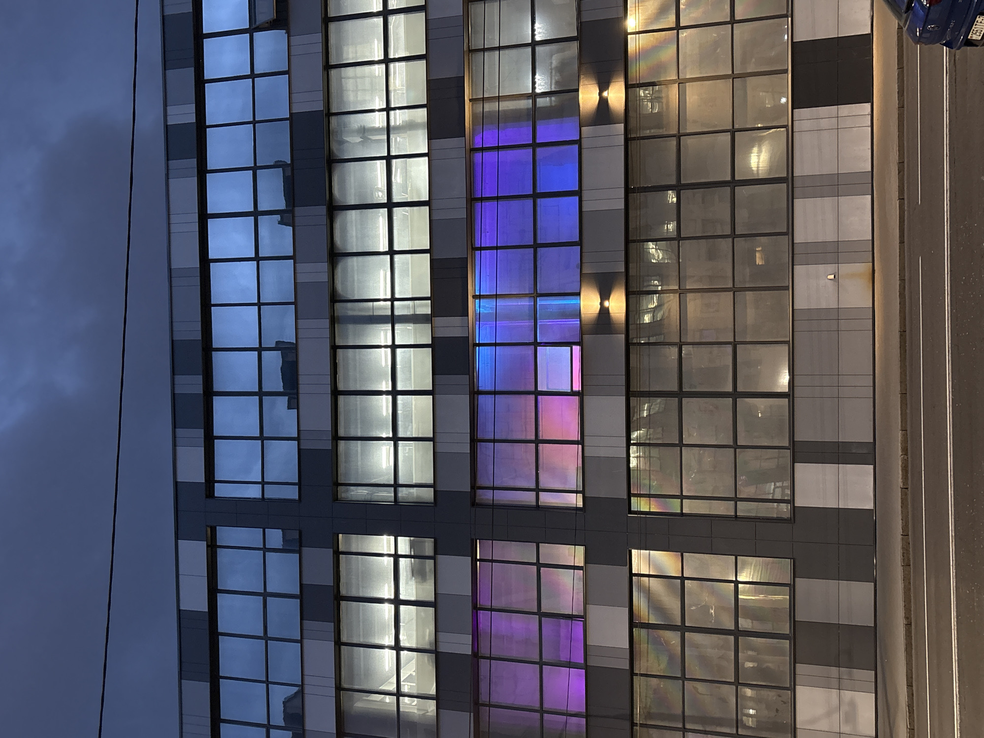 The image shows the exterior of a modern building, likely an office or commercial space, in Minsk, Belarus. The facade is dominated by a grid of windows. Some windows reflect daylight, others appear illuminated from within, suggesting different activities in the building. A column of windows features purple and pink lighting or reflections, contrasting with the more neutral tones of adjacent windows. A road is visible to the right, including the presence of a blue car. The overall environment suggests a dusk or evening setting in an urban area.