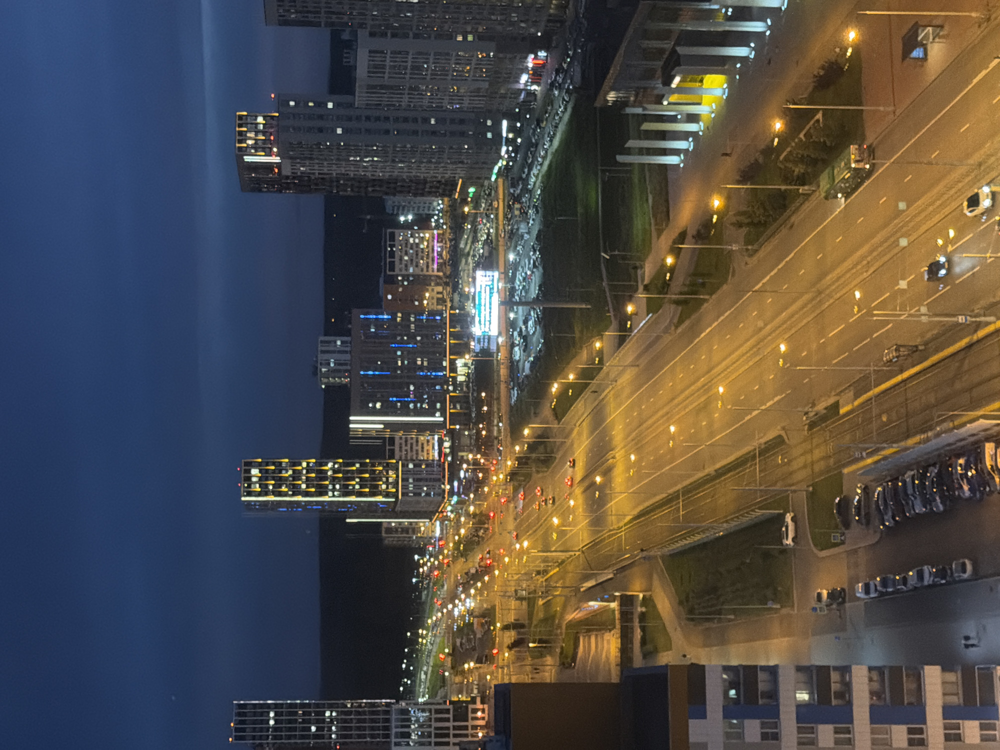 The image depicts a city scene at night, likely in an urban environment with modern architecture. A multi-lane road runs through the center of the frame, illuminated by streetlights and the headlights and taillights of numerous vehicles. Cars and what appears to be a bus are visible on the road, suggesting traffic flow. On either side of the road are buildings, including tall, illuminated skyscrapers with distinct architectural features and lighting. Some buildings have a repetitive window pattern, while others are adorned with decorative lights. In the background, a darker, possibly hilly or mountainous landscape can be seen against the night sky. The overall impression is one of a bustling city after dark, with artificial lights dominating the scene.