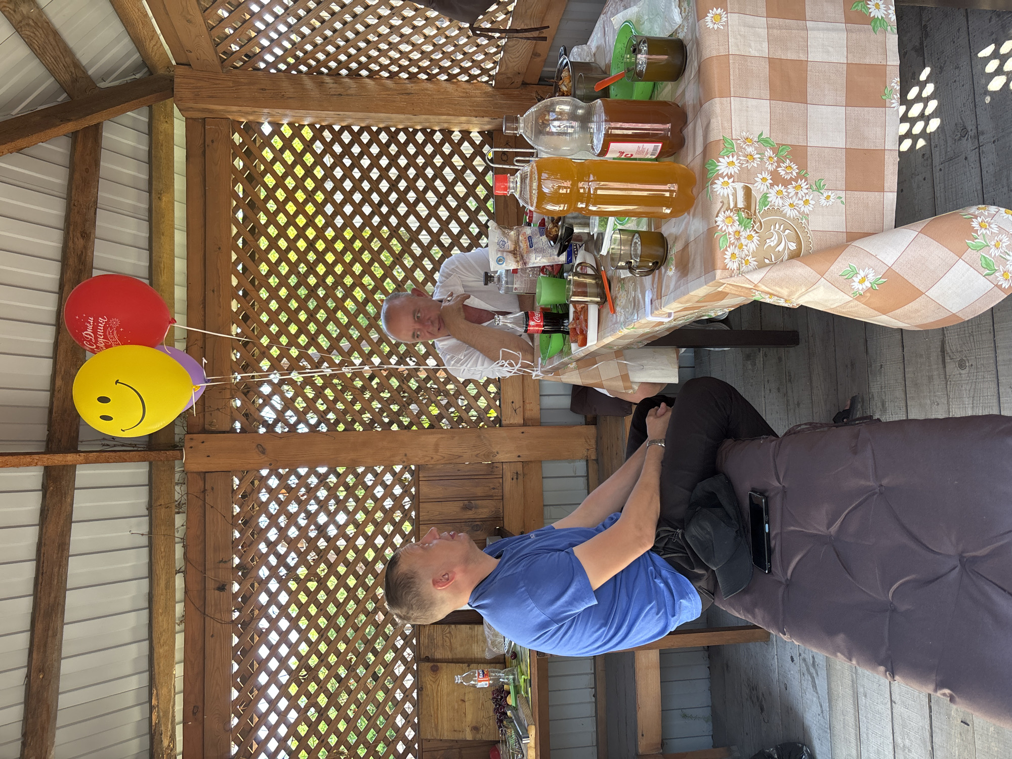 The scene is an outdoor wooden gazebo in Зелёное, Russia, constructed with stained brown lumber and lattice walls under a light gray corrugated metal roof. Two adult men are seated within the structure, engaged in an interaction.
On the left, a man with short, light brown hair and a blue short-sleeved t-shirt sits on a brown cushioned bench. He wears dark trousers and a watch on his left wrist. A black baseball cap and a black smartphone are resting on the bench beside him. He is turned towards the right, looking at the other man.
On the right, an older man with a balding head and short, light gray hair is seated at a rectangular table. He wears a white short-sleeved collared shirt with the sleeves rolled up. His right hand is near his chin, while his left arm rests on the table.
The table is covered with a light brown and white checkered tablecloth featuring a prominent daisy floral pattern. It holds an assortment of beverages and food items. There are two large plastic bottles of amber-colored liquid, one with a red cap and the other with a white and red label. A Coca-Cola glass bottle, a small plastic bottle of dark liquid, and several metal mugs and clear glasses containing dark liquids are also present. Food items include a green bowl of red contents (possibly tomatoes), a white bag, and a covered metal pot. An orange plastic spoon is visible on the tablecloth.
In the upper left portion of the image, three helium balloons are tied together: a large yellow balloon with a black smiley face, a red balloon with white Cyrillic text "С Днём Рождения" (Happy Birthday), and a partially visible purple balloon beneath them, all with white ribbons.
To the left of the man in the blue shirt, on a wooden side table, there is a metal basket containing red cherries, green vegetables (including a cucumber and possibly a bell pepper), a Fanta bottle, and another clear bottle. Green foliage is visible through the lattice work of the gazebo walls, and sunlight casts dappled shadows onto the wooden floorboards.