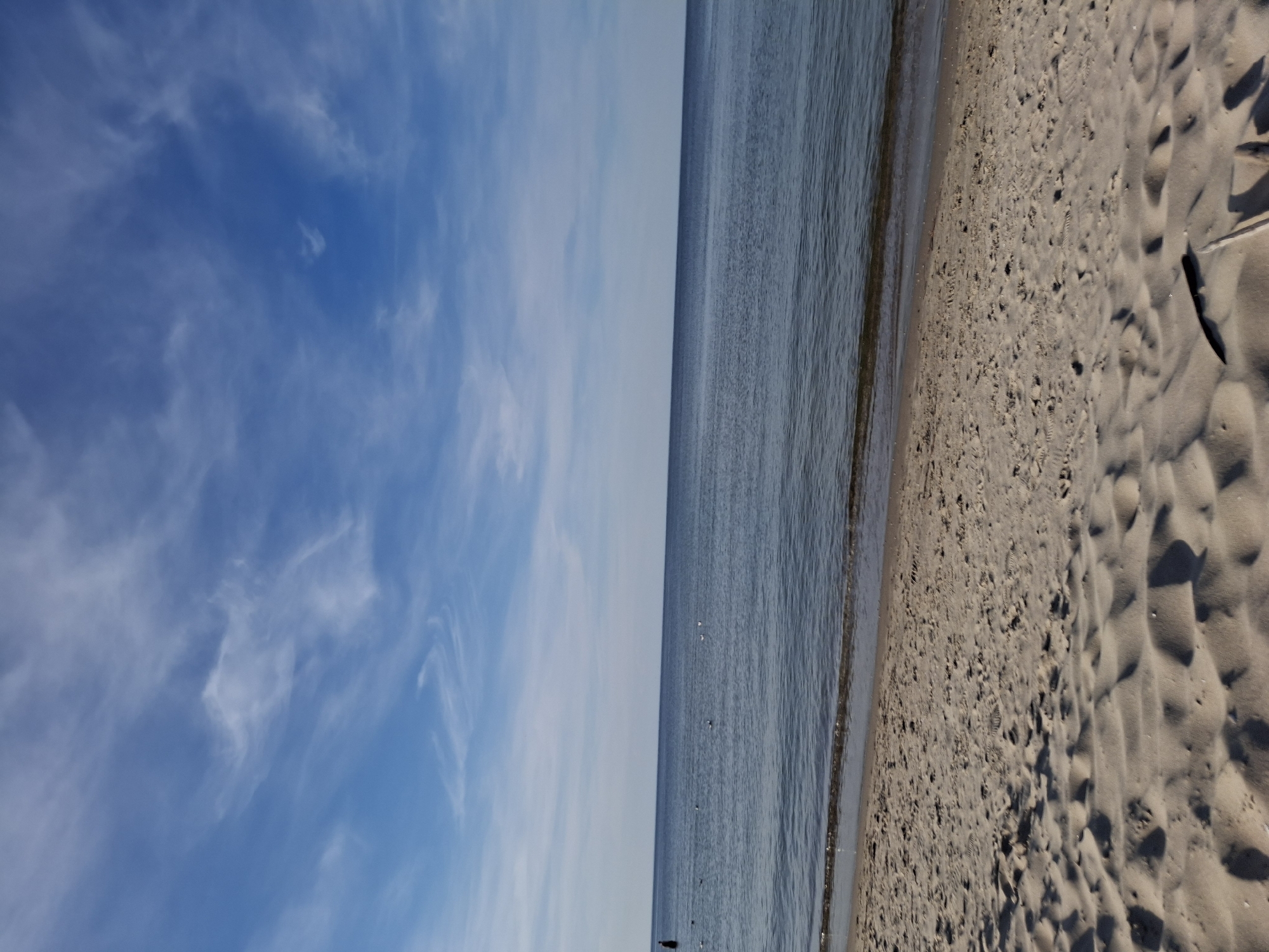 This is a photograph taken at the beach in Jantar, Poland. The image shows a sandy beach in the foreground with ripples and undulations in the sand, suggesting footprints and the wind. The sea is in the middle ground, appearing calm with gentle waves lapping at the shore. The water is a deep blue-grey. Scattered on the water are several white birds, likely seagulls. In the far distance, a dark figure, possibly a person, is visible standing in the shallow water on the left side of the frame. The sky occupies the top portion of the image and is a clear blue, dotted with wispy white clouds. The horizon line where the sea meets the sky is straight and unbroken. The overall scene depicts a peaceful and serene beach environment under a bright sky.