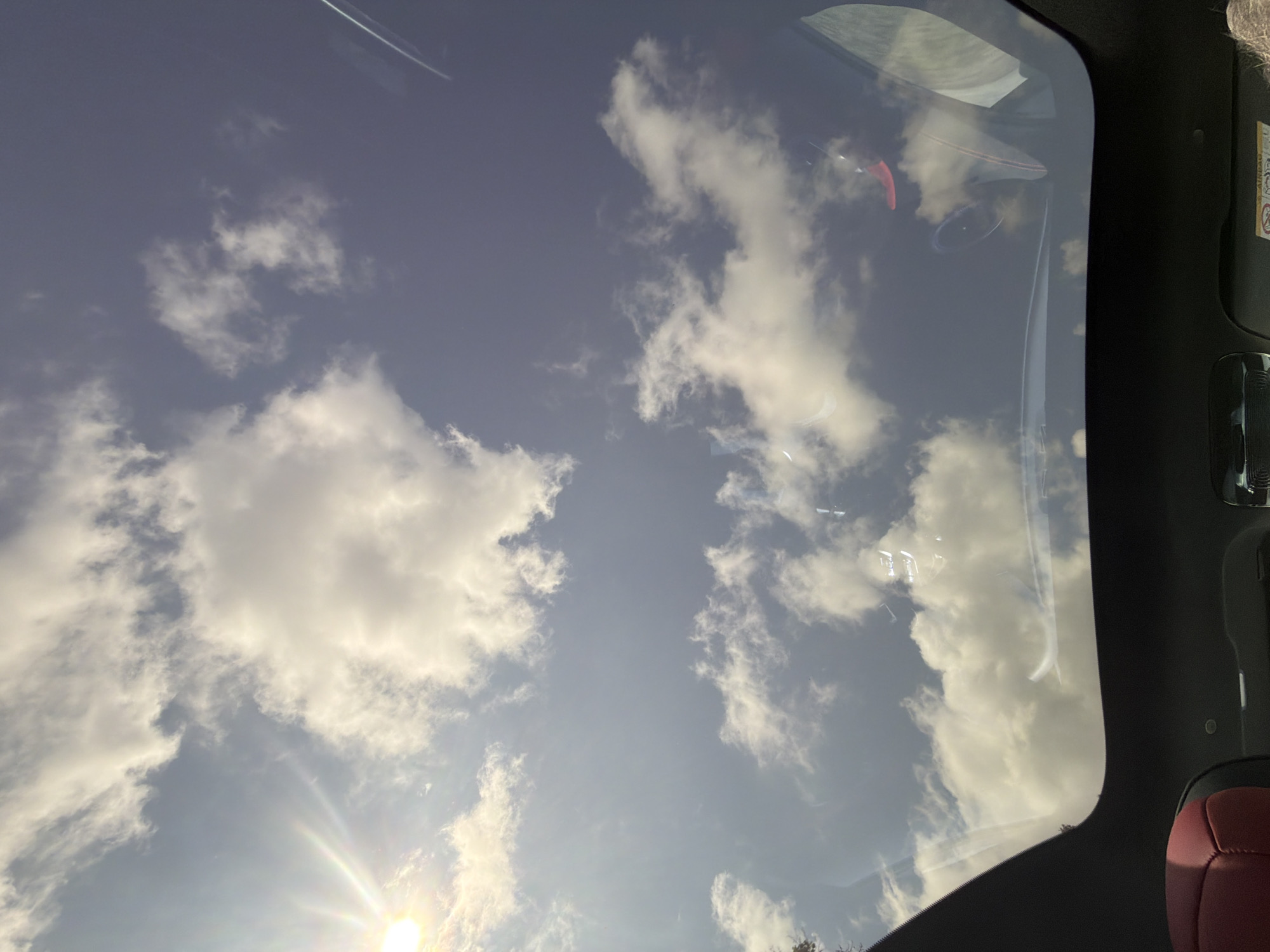 The image provides an upward view of a bright, cloudy sky as seen from inside a vehicle, likely through a panoramic sunroof or a large windshield. The sky is a clear blue, dotted with numerous white, fluffy cumulus clouds. The sun is prominent in the lower-left corner, emitting bright rays and a visible lens flare effect, indicating daytime, probably mid-morning or afternoon, with sunny weather.
The edges of the vehicle's interior frame the view, showing dark-colored trim, possibly part of the headliner or pillars. Reflections of the car's interior are visible on the glass, including vague shapes that suggest a dashboard or steering wheel. In the lower-right corner, a section of dark red or maroon upholstered car seat is visible, suggesting a modern vehicle interior. Along the upper-right side, on what appears to be an A-pillar, a white label is present with partially visible text, notably the word "AIRBAG," indicating a safety warning. The scene is static, capturing the natural beauty of the sky from a comfortable, enclosed setting, potentially during travel or while parked in Trastenik, Bulgaria, as per the provided context.