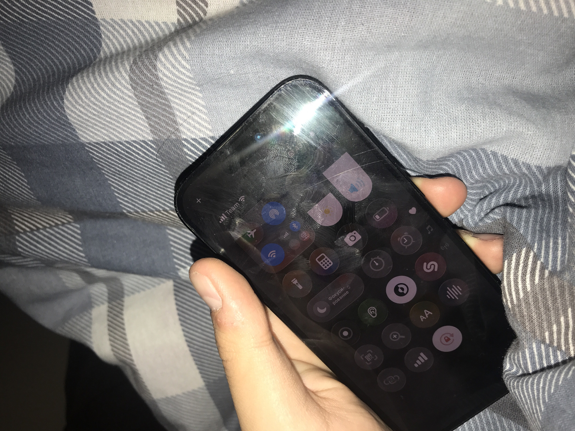 A right hand is holding a black smartphone. The screen of the smartphone is on and displaying various app icons and control settings. The phone is positioned on a checkered blue and white fabric. The location appears to be indoors. No details of the city are readily available from this photograph.