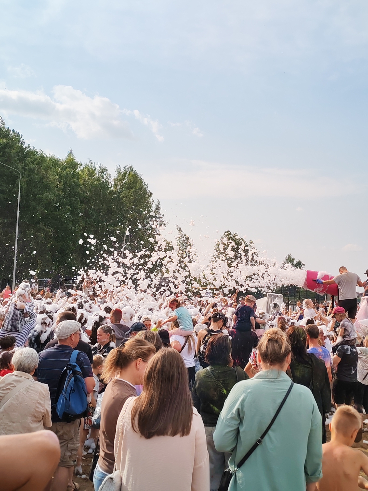 This is a full-body, vertical shot of a large crowd of people at a foam party. The scene is outdoors with a bright blue sky and white clouds above. In the background, lush green trees line the horizon. A foam machine is actively spraying a large amount of white foam into the air, which is engulfing many of the people in the crowd. Some individuals are sitting on others' shoulders, and children are visible throughout. The people are dressed in casual summer attire, and some appear to be dancing or interacting with the foam. The overall atmosphere seems festive and energetic. The scene is set in Unknown, Unknown.